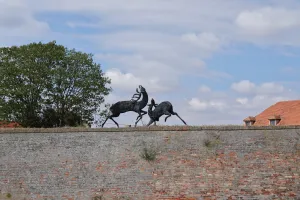 Bronze sculpture of two deer fighting or playing on top of a brick wall, with a tree and a building in the background under a cloudy sky.