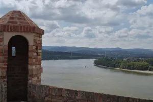 A brick lookout tower from the Petrovaradin Fortress in Novi Sad, Serbia, with the wide Danube River and a distant bridge and cityscape under a cloudy sky.