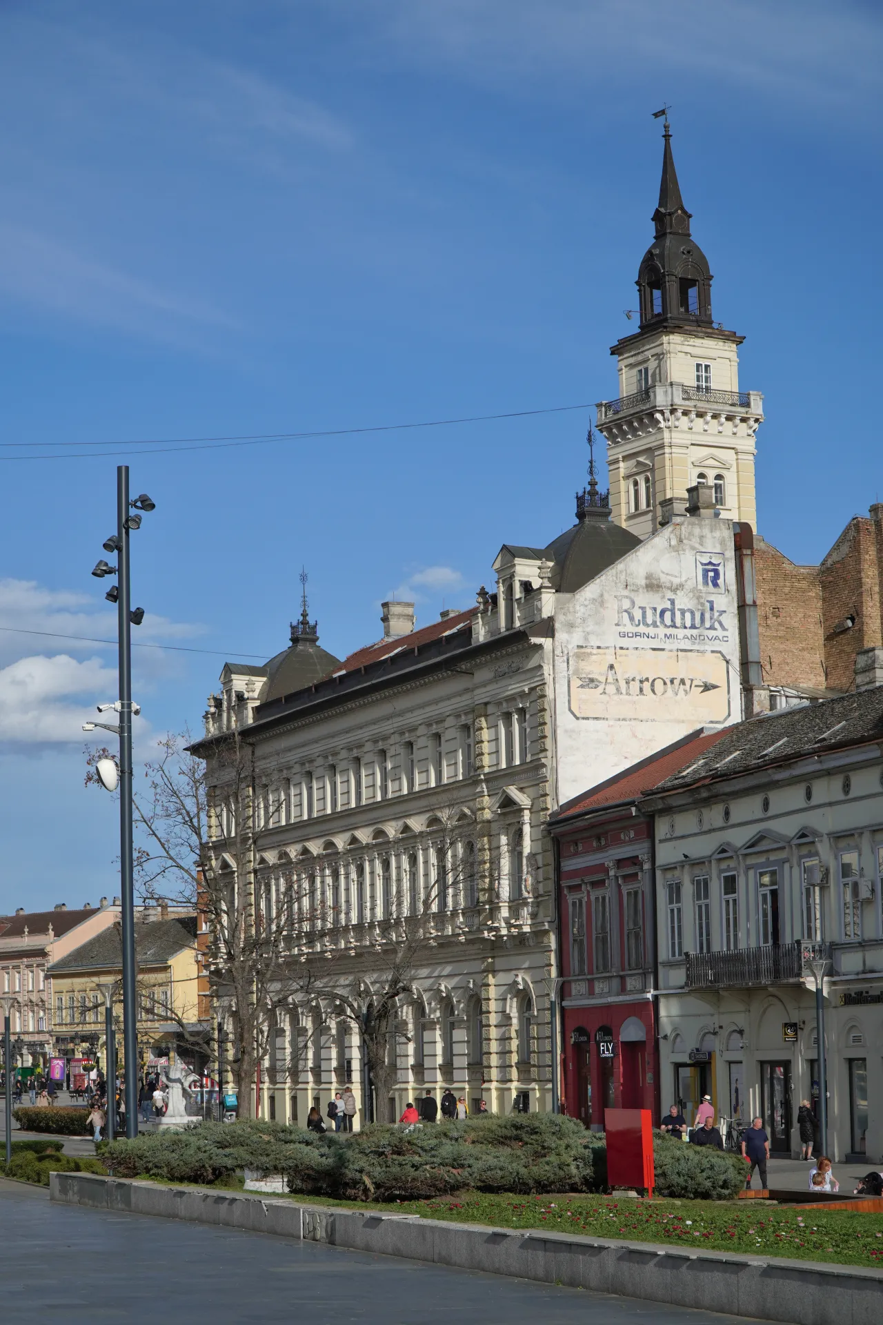 A sunny day in Liberty Square, Novi Sad, showing the City Hall tower, historical buildings, green spaces, and people enjoying the square.