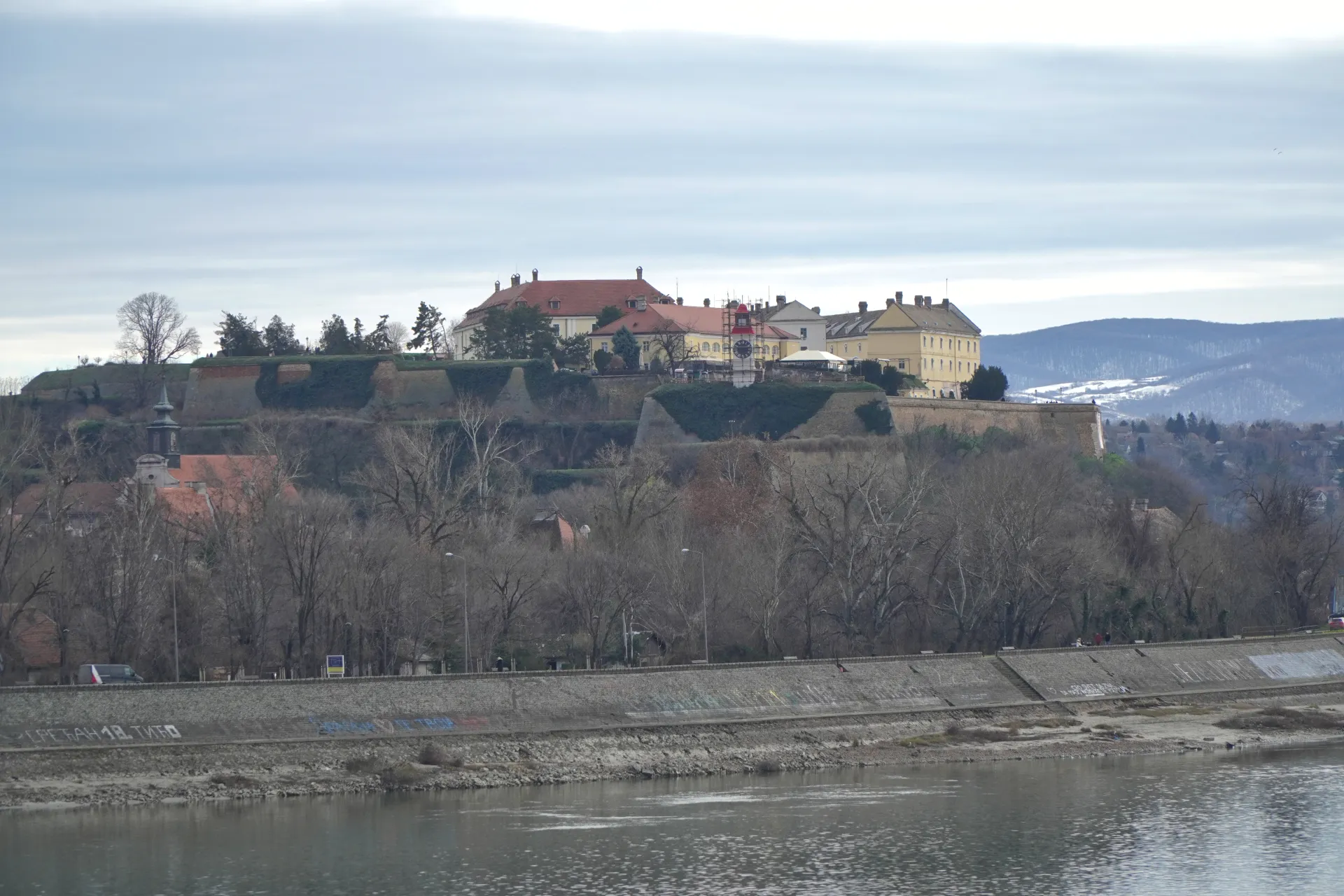 Petrovaradin Fortress perched on a hill overlooking the Danube River in Novi Sad, Serbia, on a cloudy winter day, with bare trees in the foreground and distant hills.