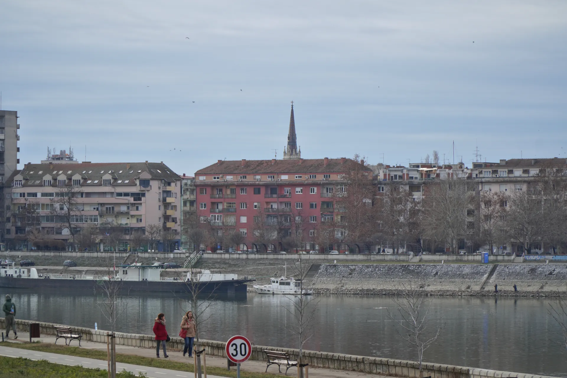 Prikaz reke Dunav u Novom Sadu, Srbija, sa stambenim zgradama duž obale i brodovima na vodi pod oblačnim nebom.