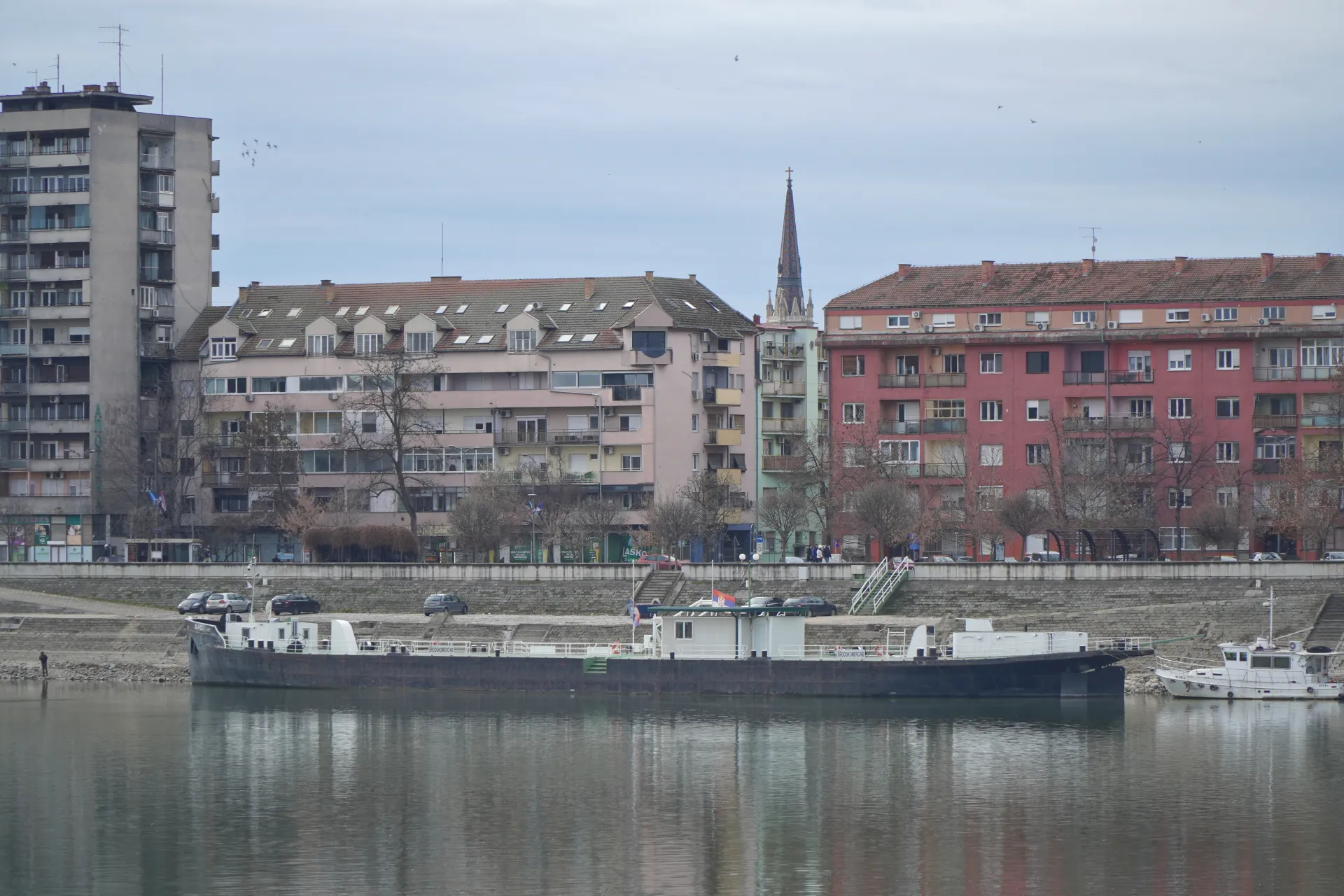 Stambene zgrade i crkveni toranj stoje na obali reke Dunav u Novom Sadu, Srbija, sa baržom i manjim brodom usidrenim u vodi.