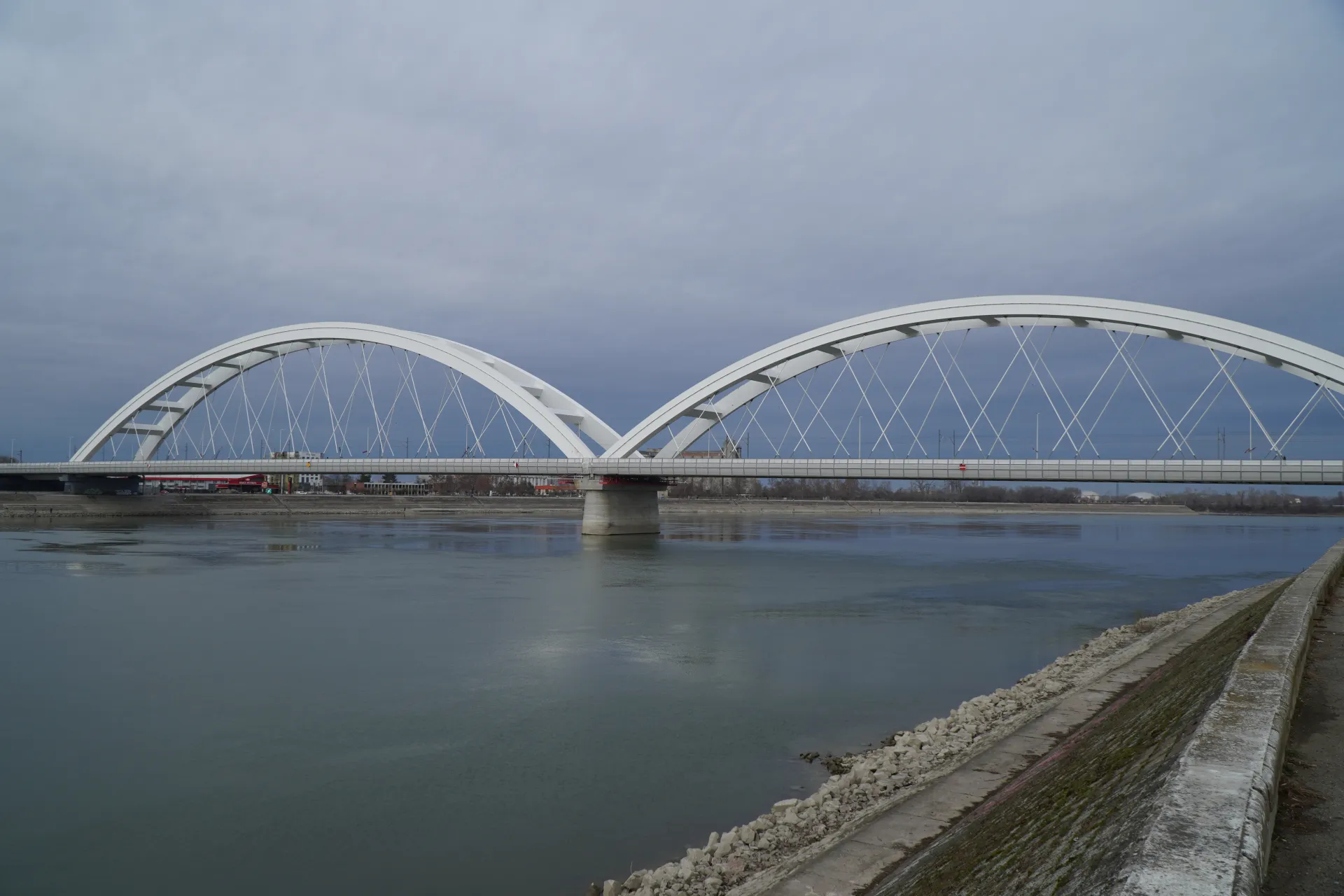 A wide shot of the Novi Sad Arch Bridge with its white double arches, spanning the Danube River on a cloudy day. The river's surface is calm, reflecting the overcast sky. The embankment is reinforced with stone and concrete.