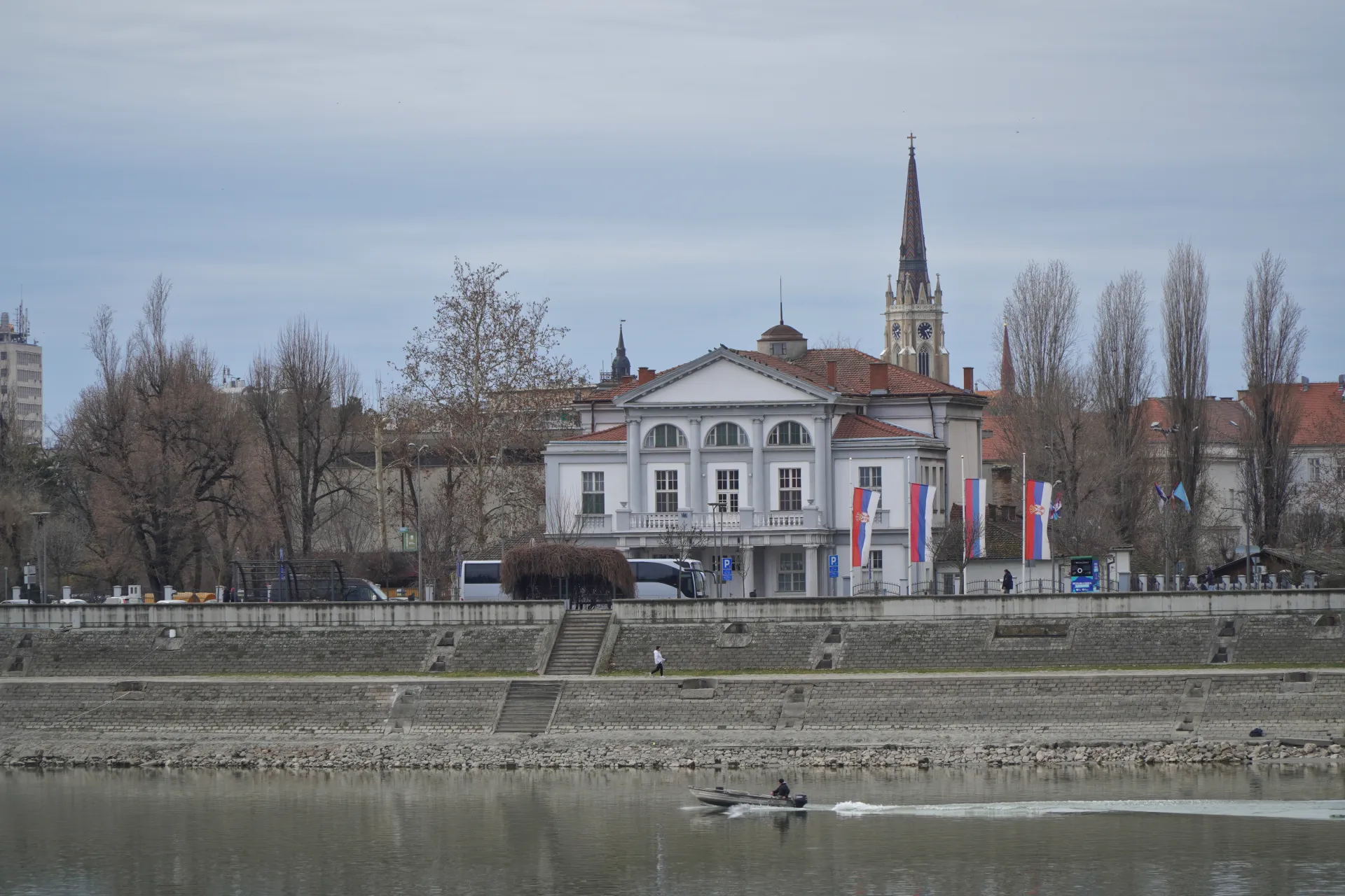Pogled na kej pored Dunava u Novom Sadu, Srbija, sa belom zgradom, srpskim zastavama, zvonikom i čamcem na reci.