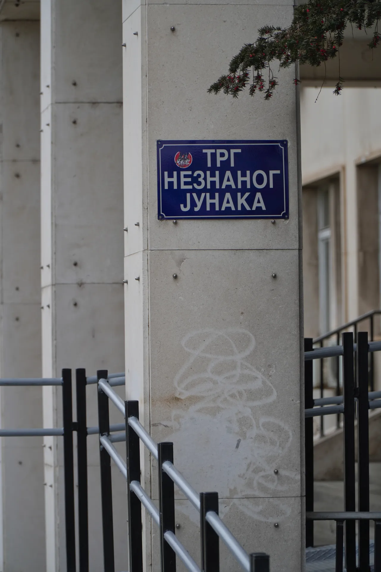 Синий уличный знак в Нови-Саде, Сербия, с надписью 'TRG NEZNANOG JUNAKA' (Площадь Неизвестного Героя), установленный на бетонной колонне с гербом Сербии. На заднем плане видны металлические перила и часть здания.