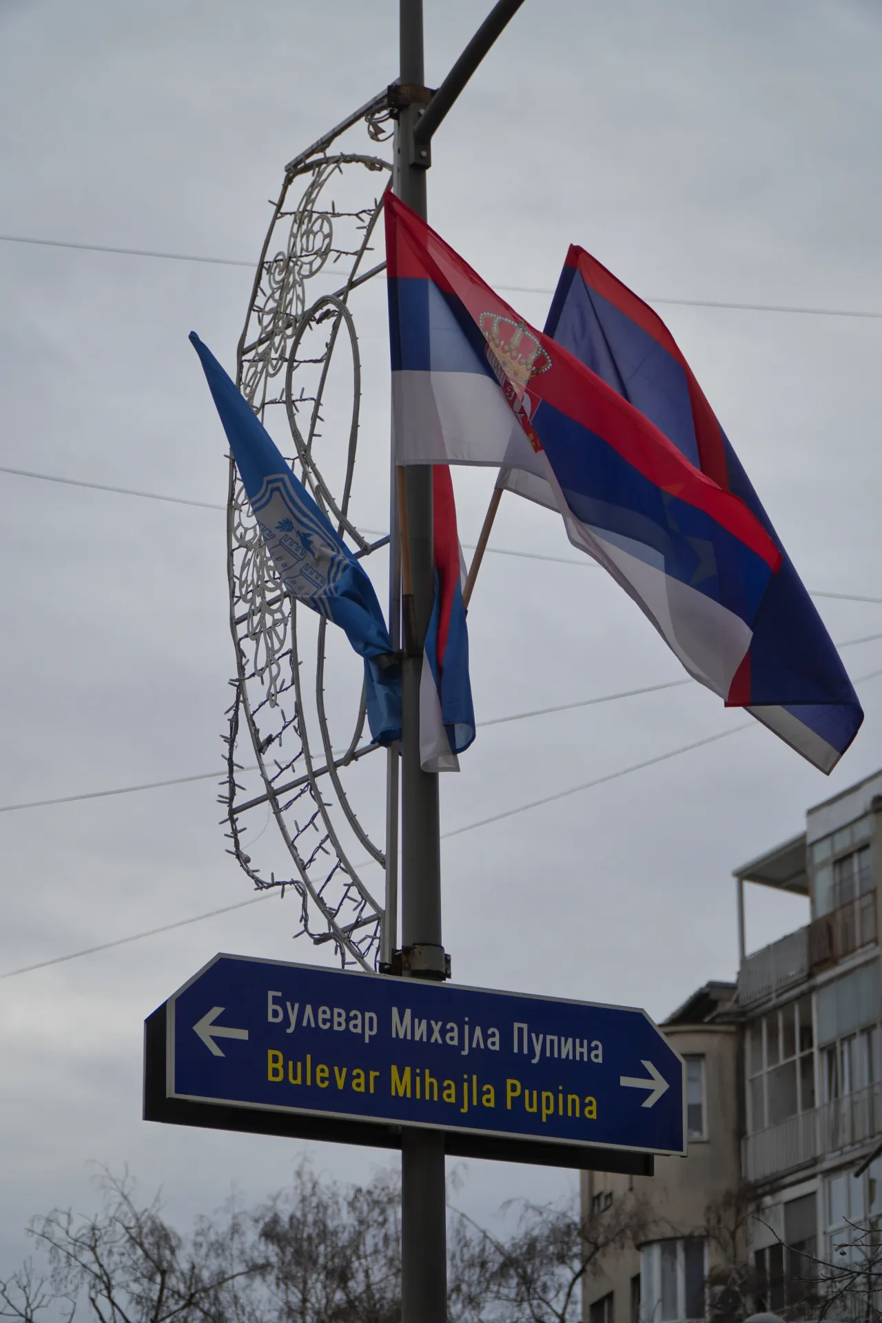 Saobraćajni znak koji pokazuje ka Bulevaru Mihajla Pupina u Novom Sadu, Srbija, sa srpskim zastavama i ukrasnom svetlosnom konstrukcijom na stubu.