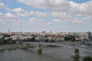 A wide shot of Novi Sad, Serbia, with the Danube River in the foreground and the city's buildings and a bridge under a sky with clouds.