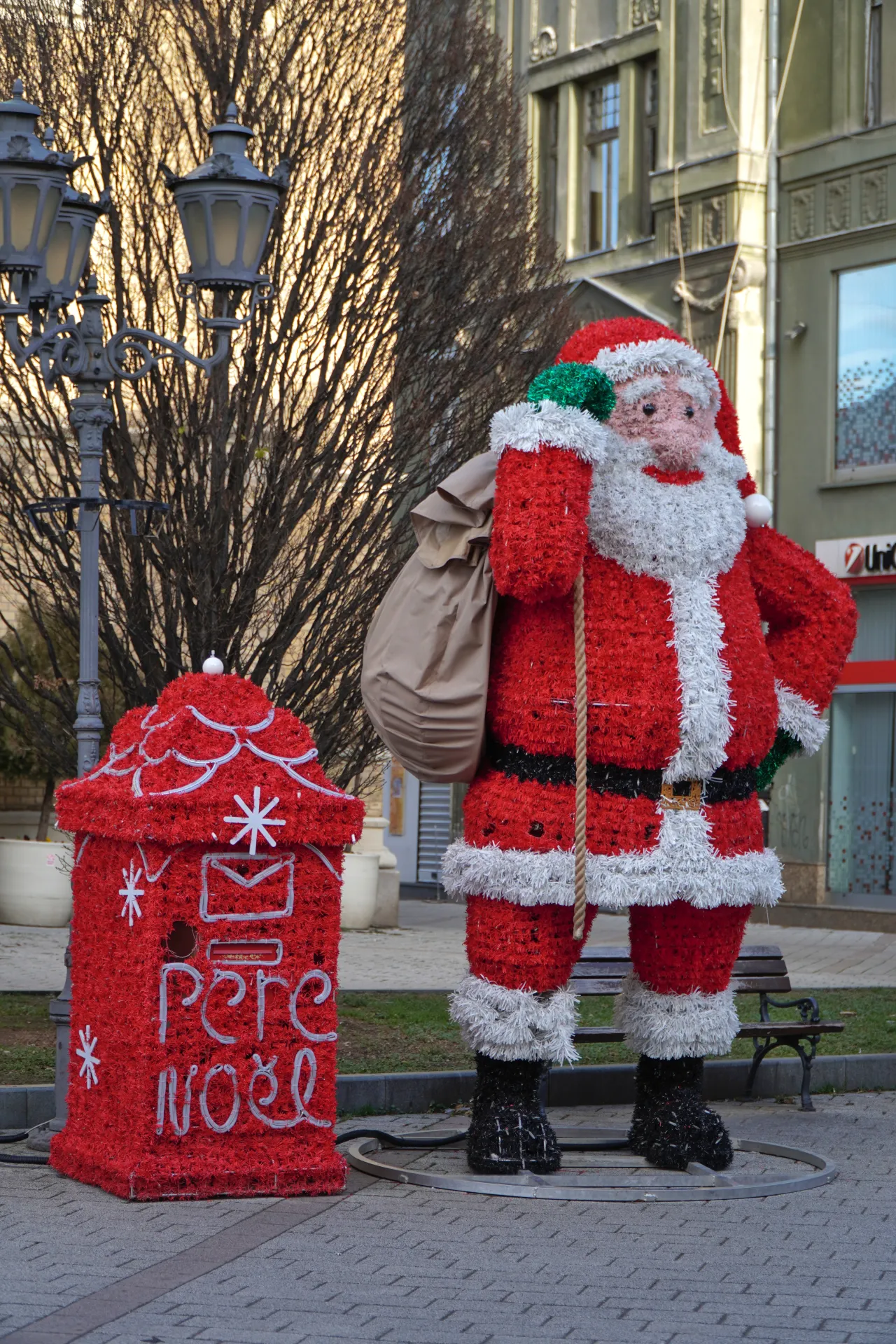 A large, fluffy Santa Claus figure stands next to a bright red post box decorated with 'Père Noël' written in white lights, situated on a paved city square in Novi Sad, Serbia, with bare trees and buildings in the background.
