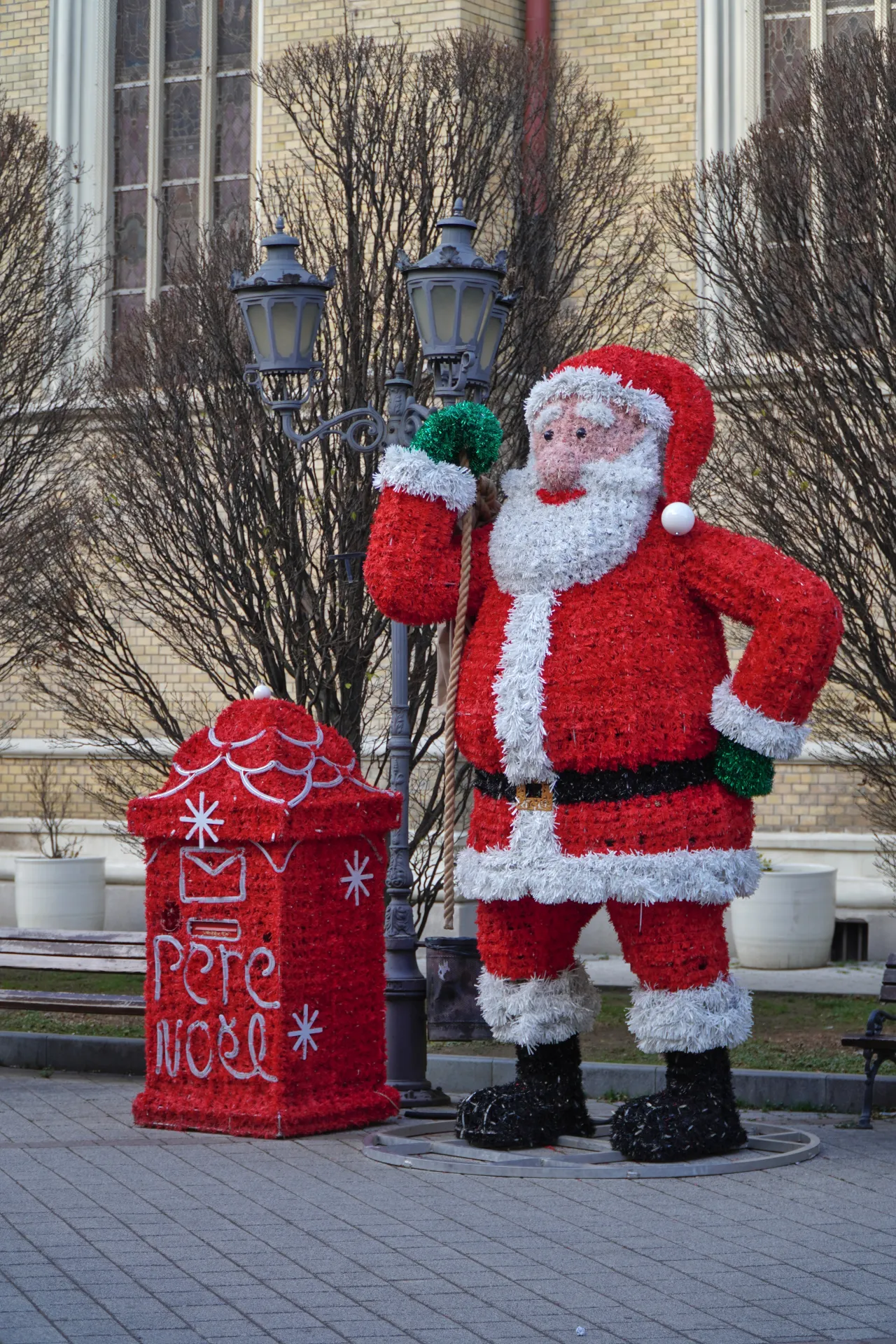 A large, illuminated Santa Claus figure and a red mailbox decorated with 'Pere Noel' and snowflakes, placed in an outdoor area in Novi Sad, Serbia during the holiday season.