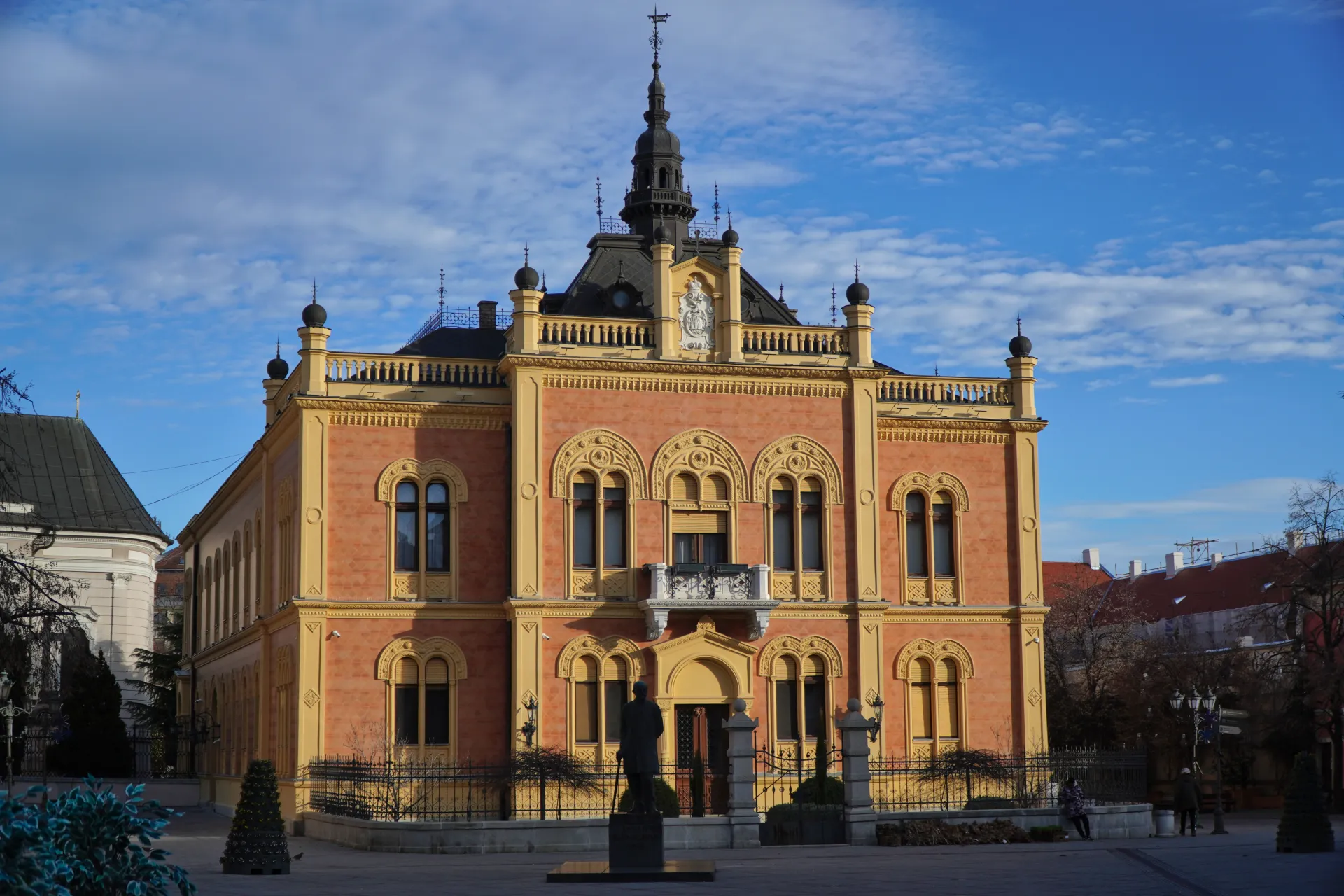 Zgrada Biblioteke Matice srpske u Novom Sadu, Srbija, sa plavim nebom i rasutim oblacima u pozadini. Zgrada poseduje istaknut toranj, ukrašene prozore i statuu u prvom planu.