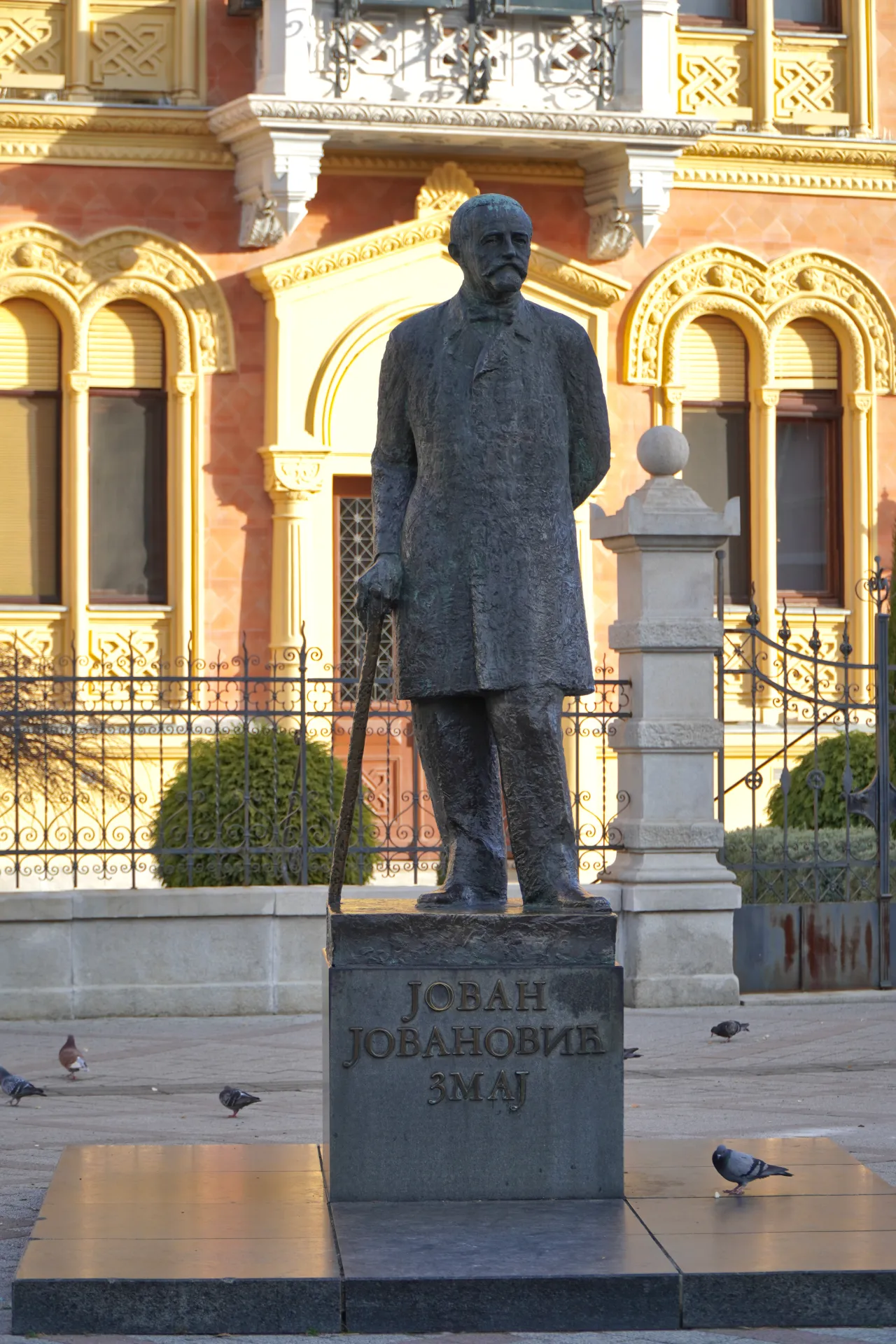 Bronzana statua Jovana Jovanovića Zmaja, srpskog pesnika, koji drži štap, stoji na postolju ispred ukrašene zgrade u Novom Sadu, Srbija. Golubovi su rasuti po pločniku.