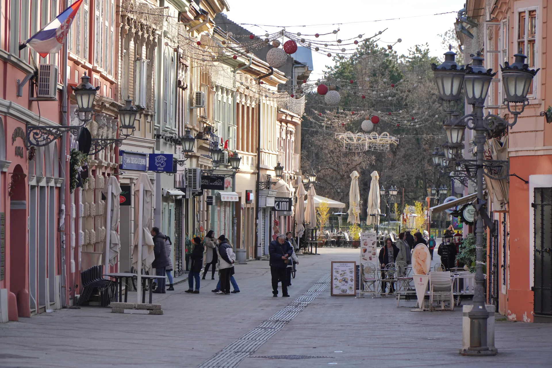 Sunčan dan u Dunavska ulici u Novom Sadu, Srbija, sa šarenim zgradama, prodavnicama, baštama kafića i prazničnim ukrasima okačenim preko pešačke staze.