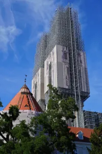 Zagreb Cathedral partially covered by scaffolding and renovation tarpaulins, with a red-roofed building in the foreground under a blue sky.