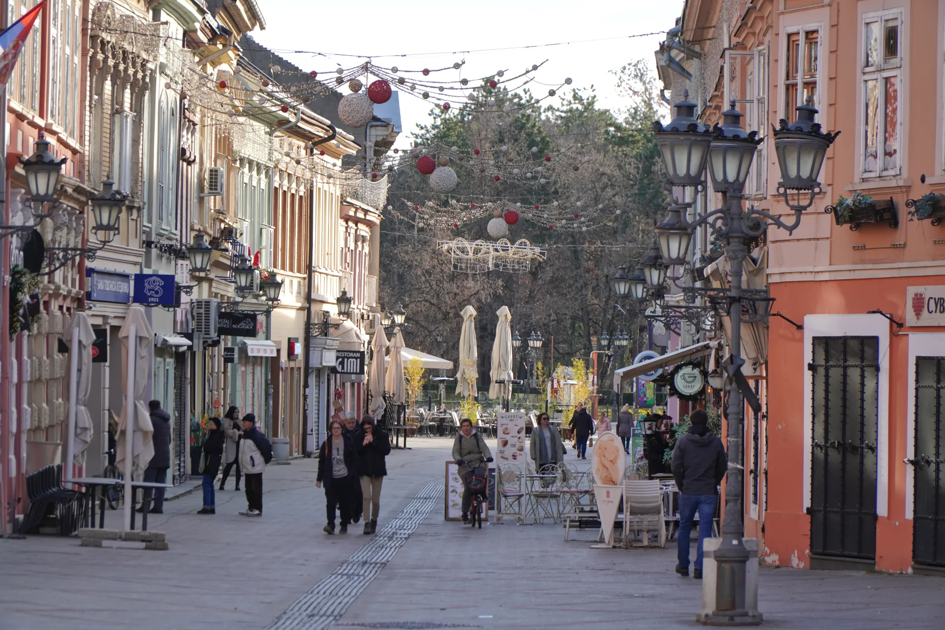 A view down a bustling pedestrian street in Novi Sad, Serbia, with people walking, shops, cafes, and decorative lights strung overhead.