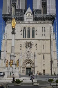 A view of the Zagreb Cathedral with its iconic golden Statue of the Virgin Mary in the foreground, set against a clear blue sky. Scaffolding is visible on parts of the cathedral's towers, indicating ongoing work.