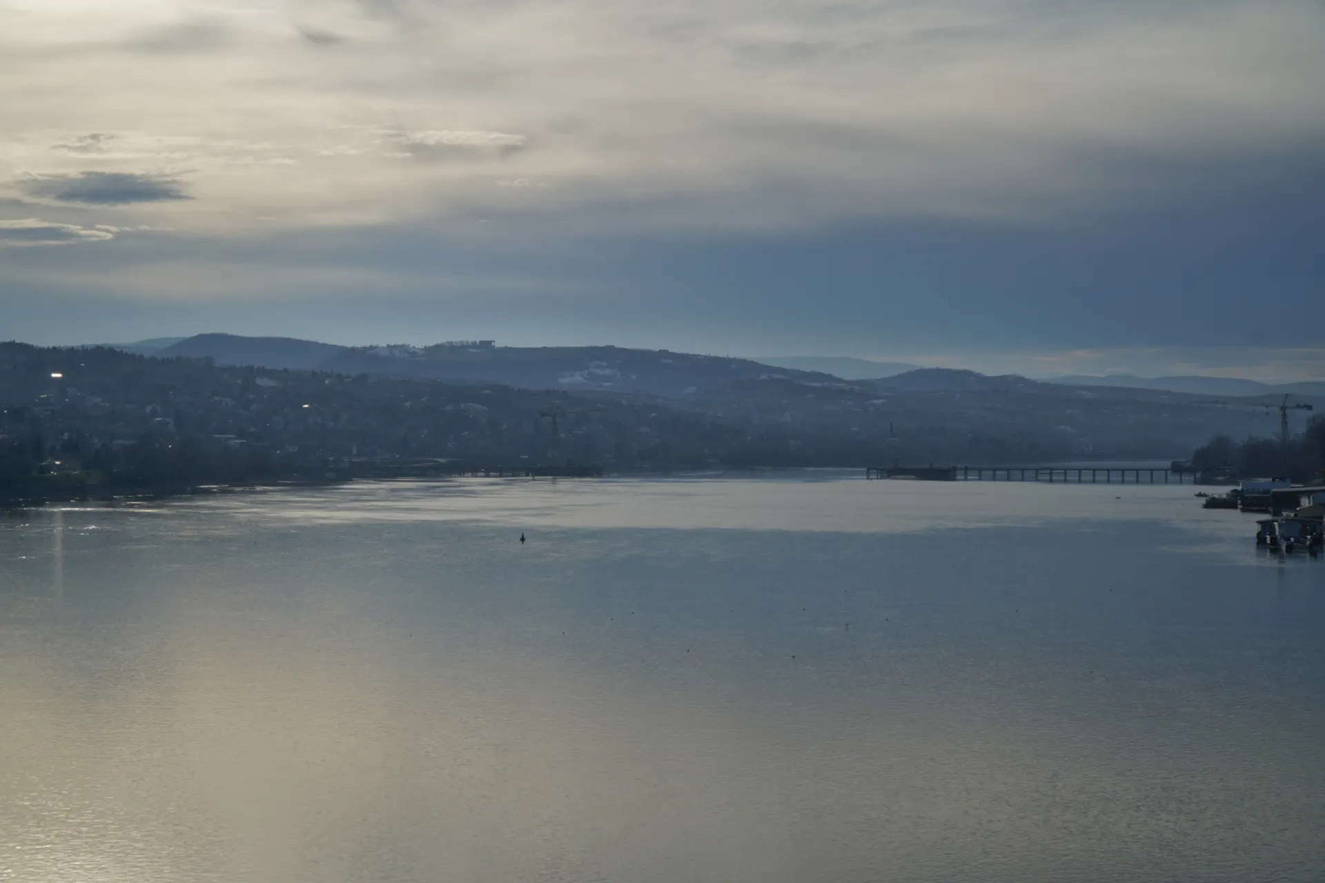 A wide shot of the Danube River flowing through Novi Sad, Serbia, with the city's buildings and hills visible in the background under a cloudy sky.