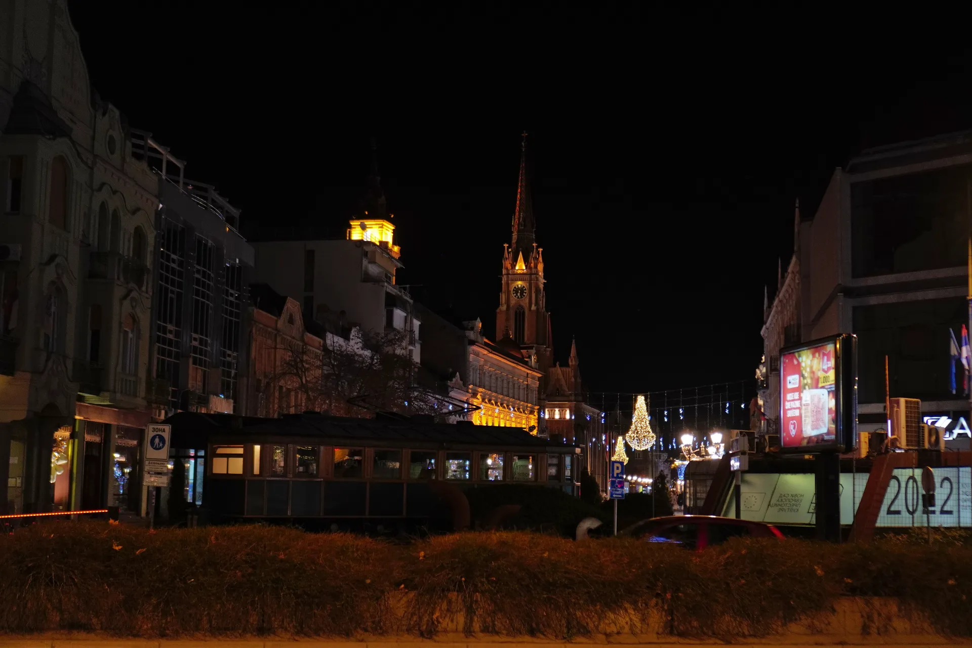 Nighttime view of Novi Sad city center with a prominent church spire, illuminated buildings, streetlights, decorative lights, and a vintage tram on the street.