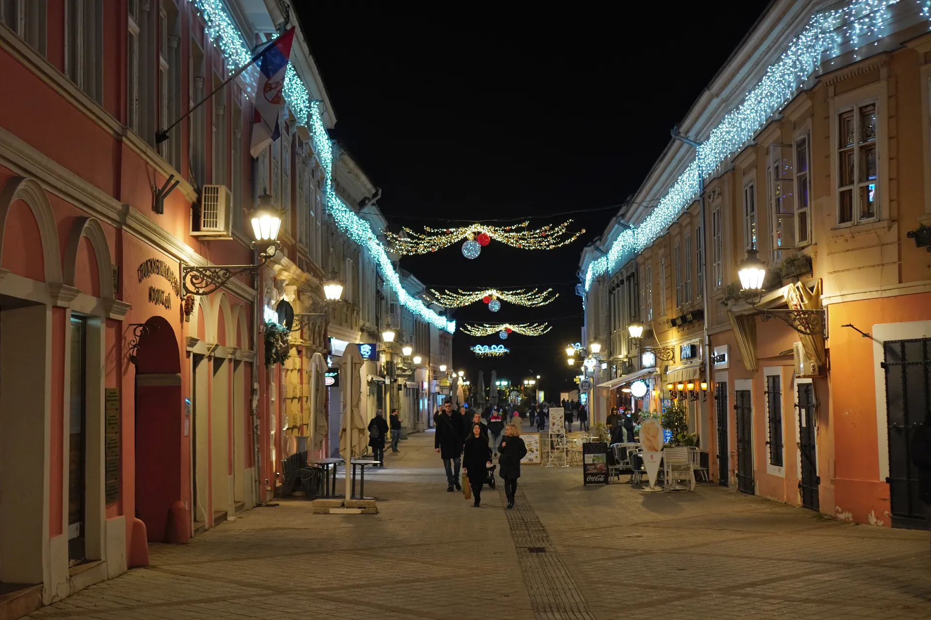 Živopisna pešačka ulica u Novom Sadu, Srbija, noću, ukrašena brojnim nizovima svečanih svetala koja se protežu ulicom, osvetljavajući fasade starih zgrada i ljude koji prolaze ispod.