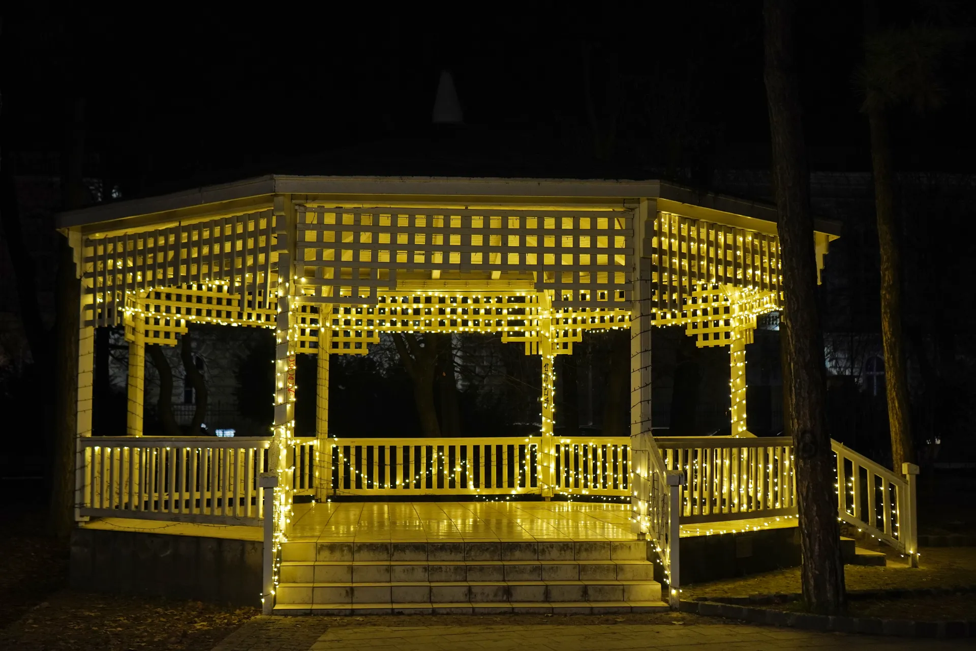 A hexagonal white gazebo adorned with warm yellow string lights, standing under the night sky in Novi Sad, Serbia. Steps lead up to the entrance, and trees are visible behind the structure.