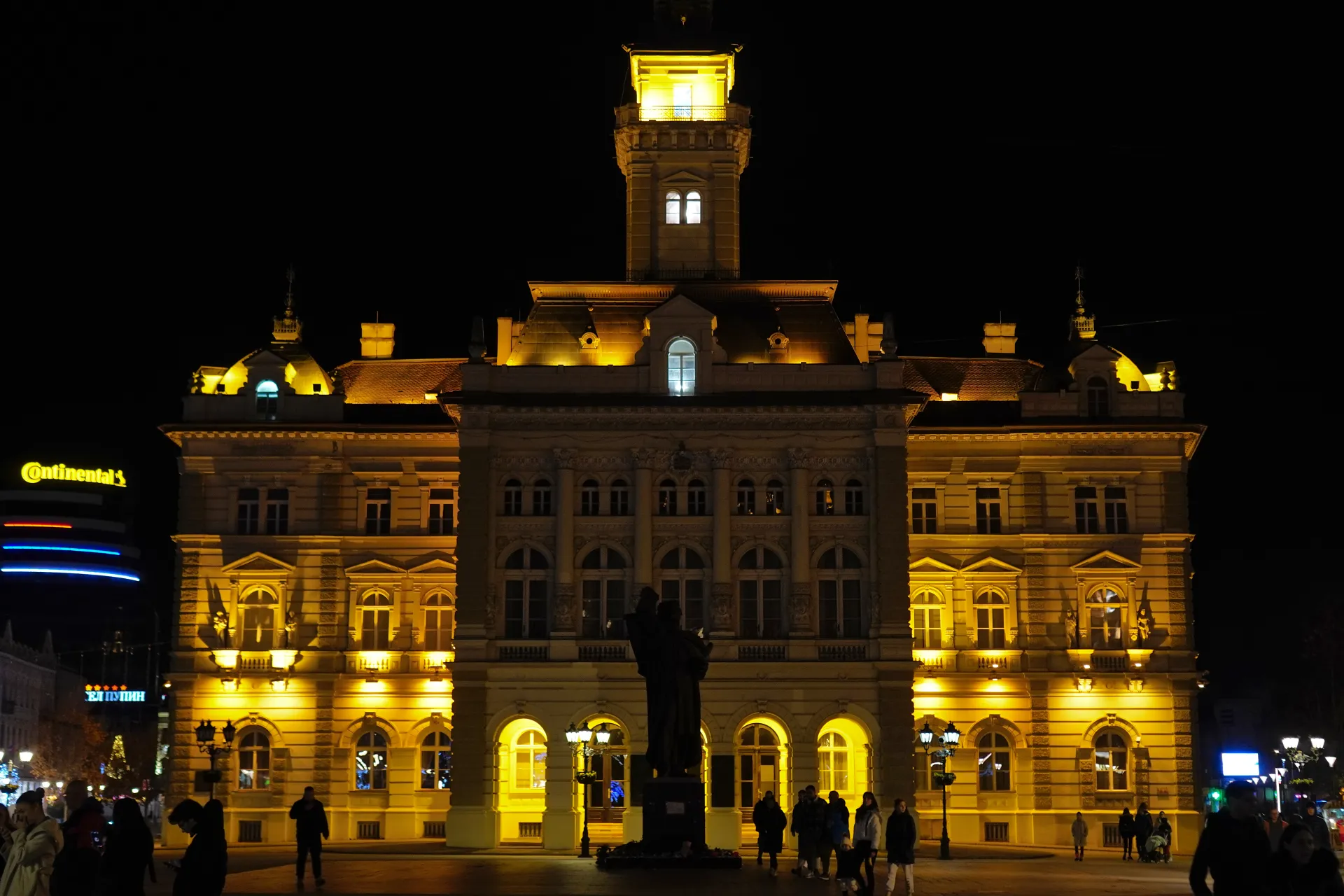 A grand building, the City Hall of Novi Sad, Serbia, is illuminated with bright yellow lights at night. The architecture features multiple windows, decorative elements, and a prominent tower. People are visible in the foreground, suggesting a public space.