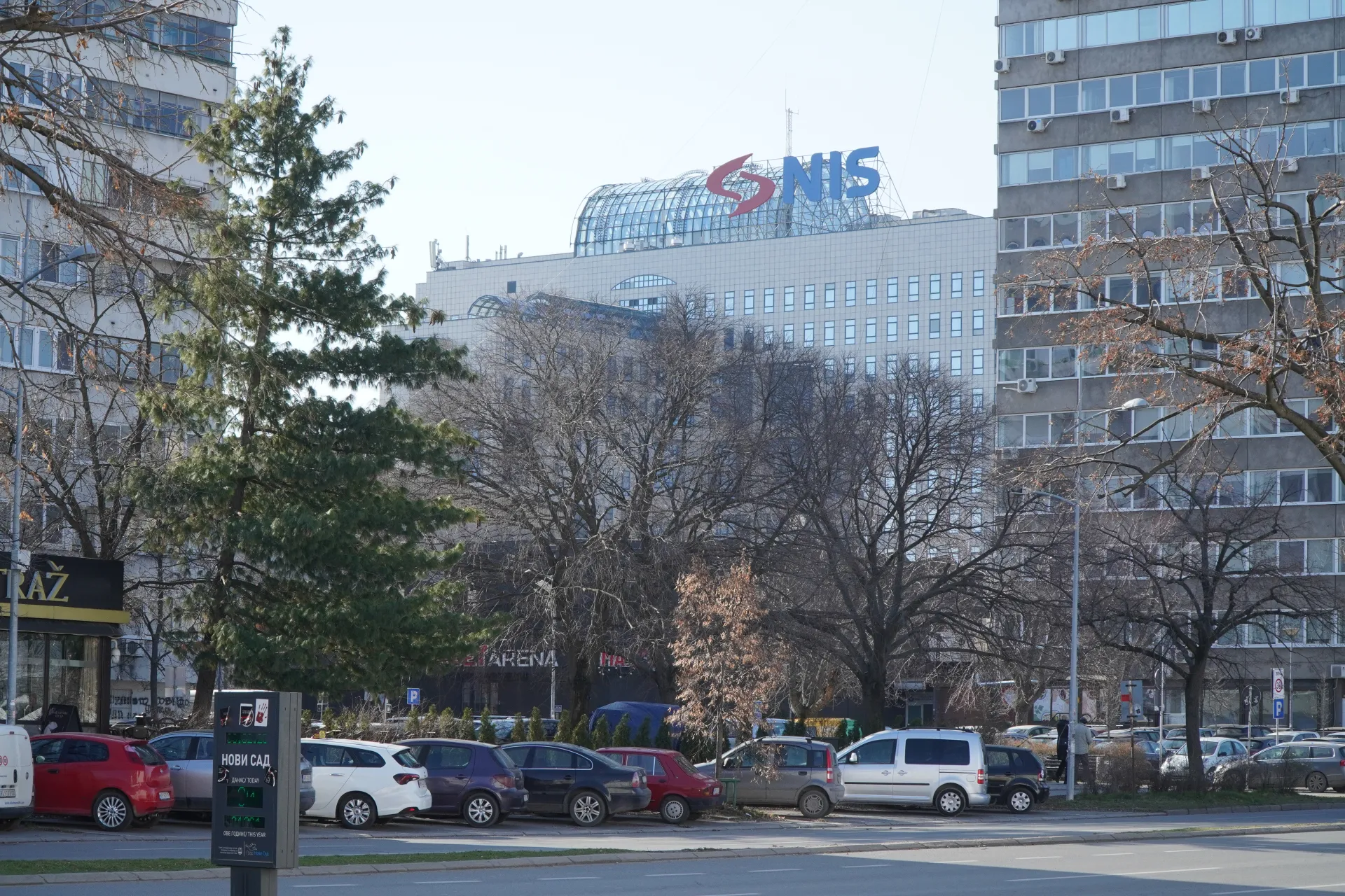 The NIS company logo is visible on top of a modern office building in Novi Sad, Serbia, with bare trees and parked cars in the foreground.