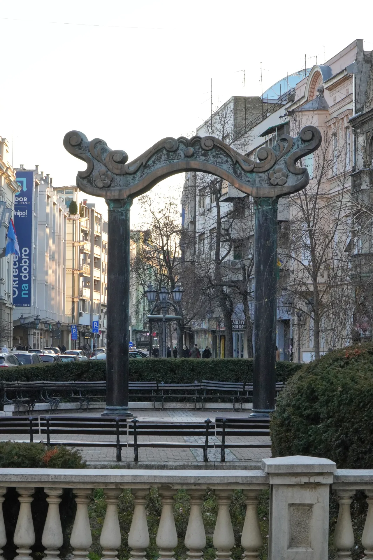Ukrasni bronzani luk sa zamršenim rezbarijama stoji na gradskom trgu u Novom Sadu, Srbija, sa zgradama i ljudima u pozadini.