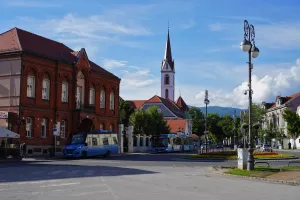 A bustling city square in Zagreb, Croatia, with a prominent red brick building, a white church tower with a spire, and blue buses on the road under a bright, cloudy sky.