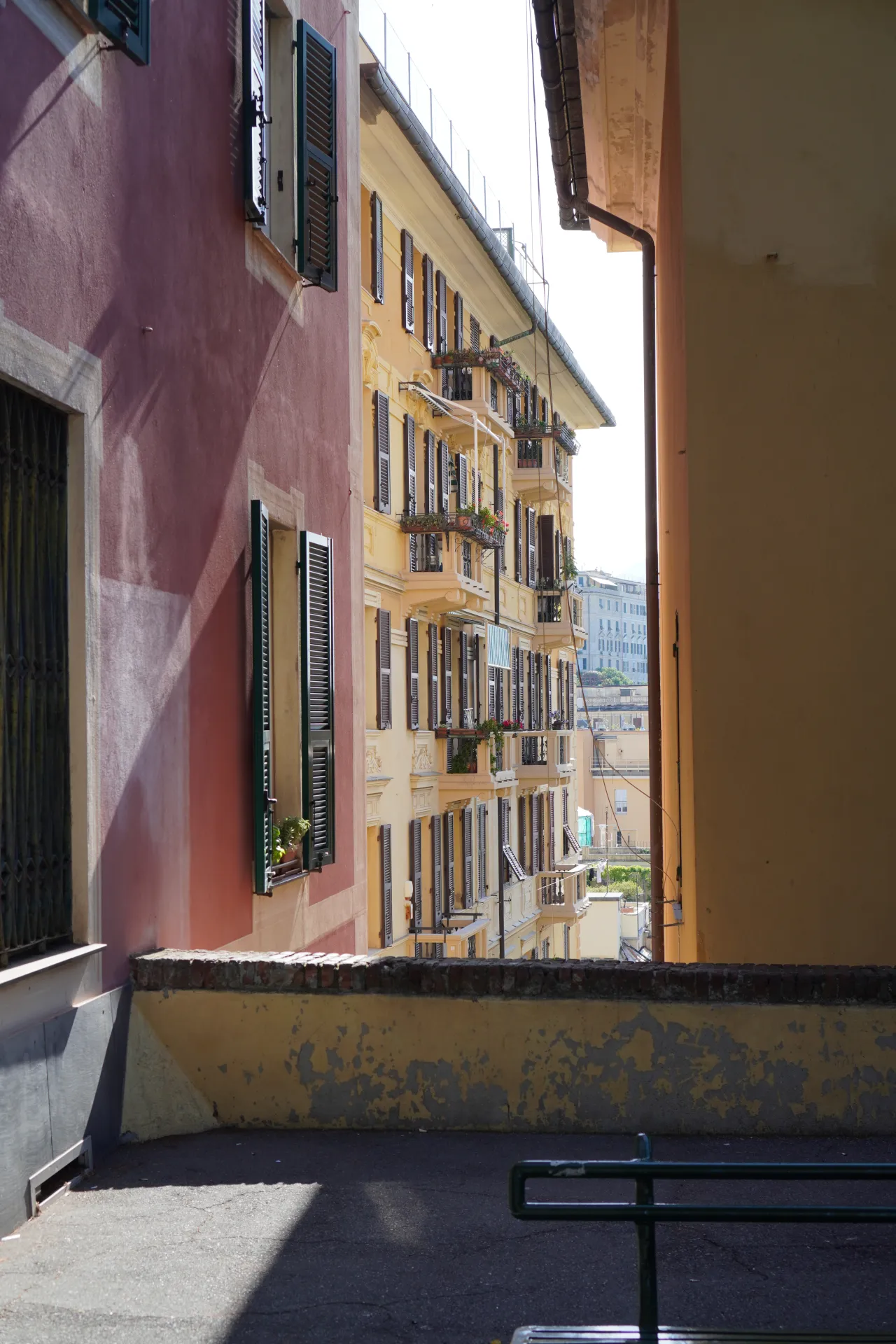 A narrow street in Genoa's old town, with a pink building on the left and a yellow building with balconies on the right, bathed in sunlight.