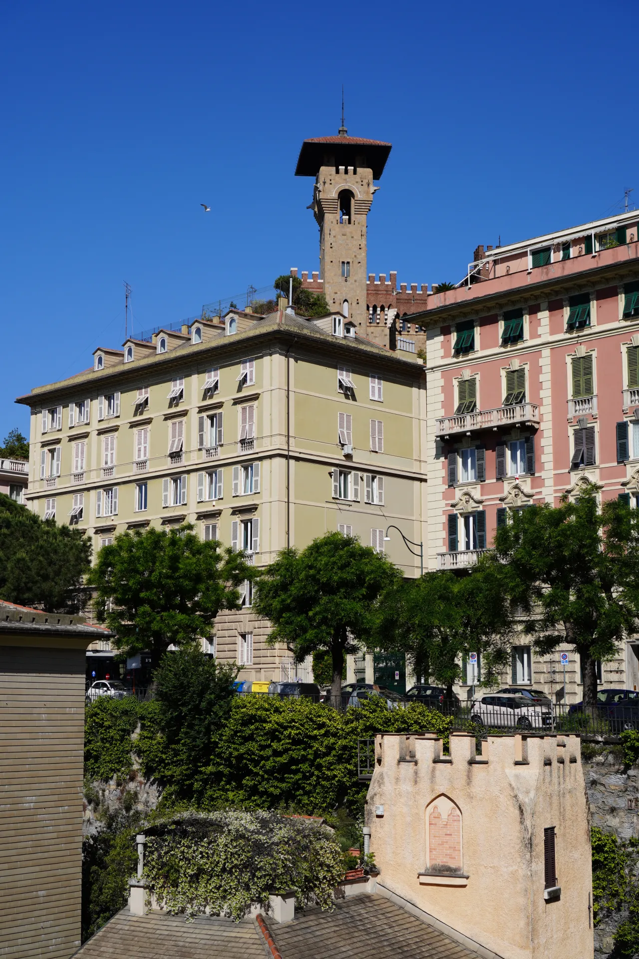 A view of Genoa, Italy, with pastel-colored buildings and a medieval-style tower under a clear blue sky, surrounded by green trees.