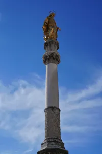 A tall stone column featuring detailed carvings, topped with a golden statue of the Virgin Mary with a halo, under a bright blue sky with scattered clouds.