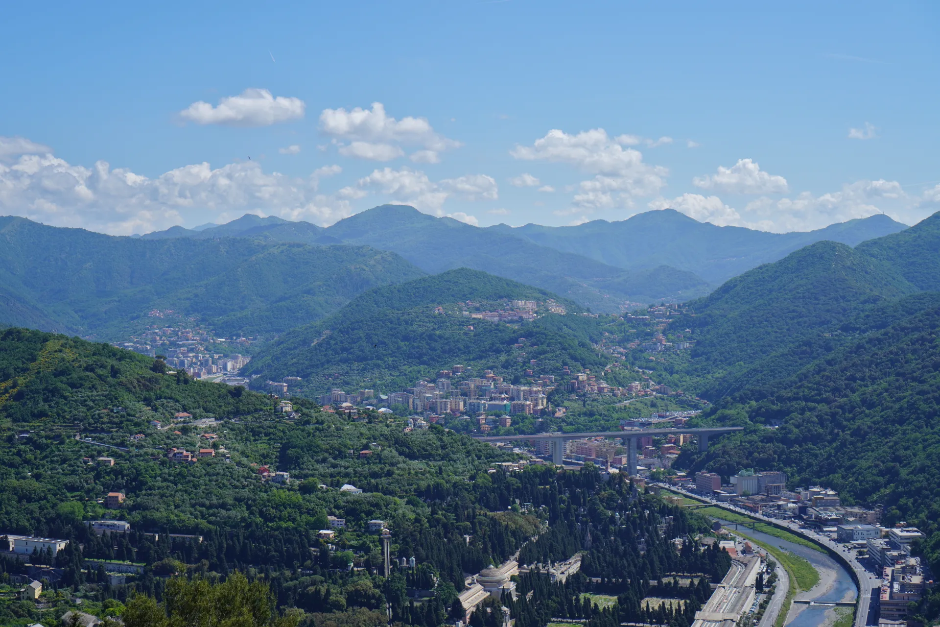 An expansive aerial view of Genoa, Italy, revealing a city built within steep, verdant valleys. Numerous buildings and residential areas are visible on the slopes and in the valley floor, interspersed with trees. A river winds through the urban landscape, and a large highway overpass crosses the scene. Distant blue-hued mountains form the backdrop under a sky dotted with white clouds.