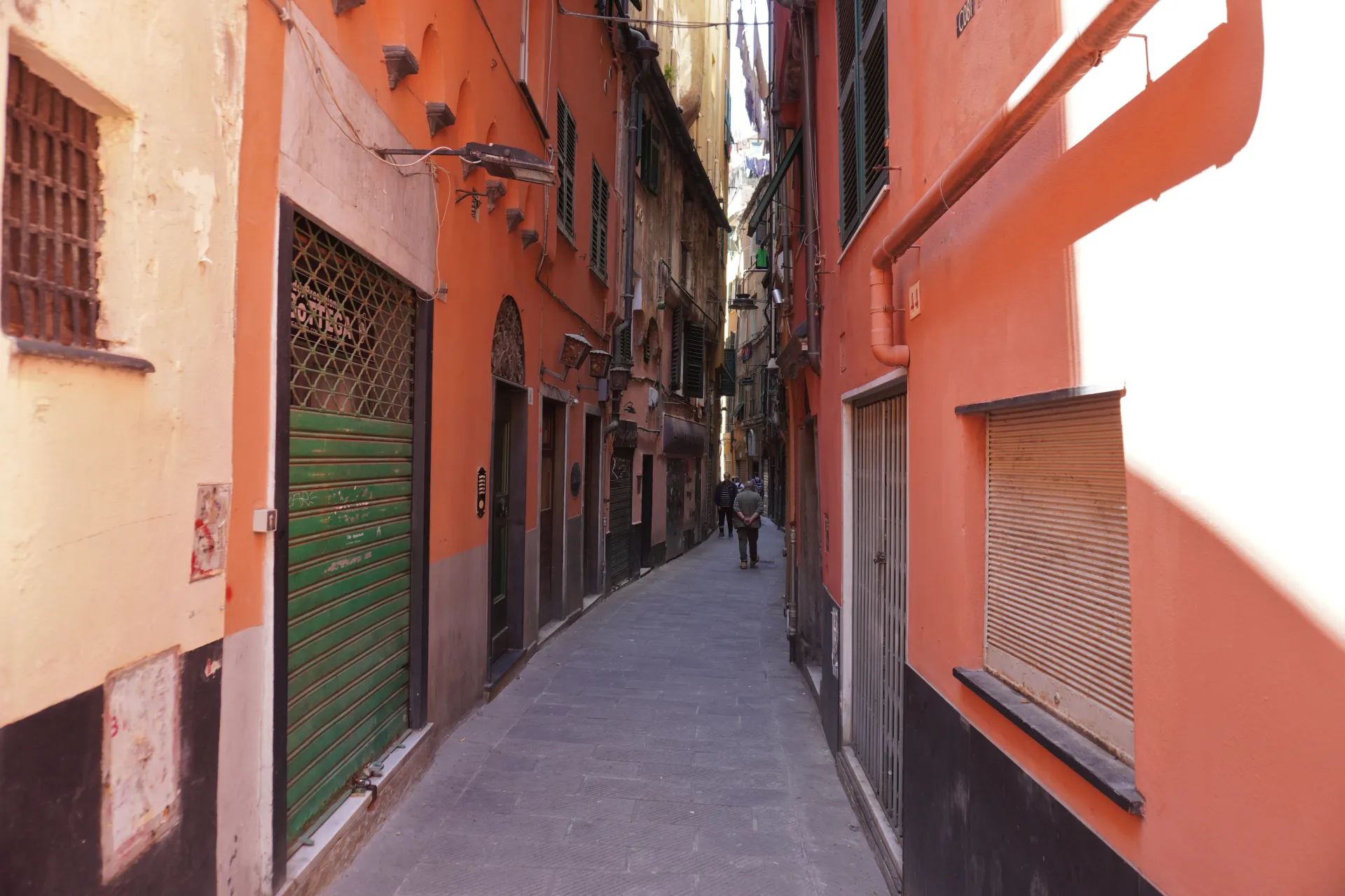 A man walks down a narrow, colorful alleyway in Genoa, Italy, with old buildings and closed shutters.