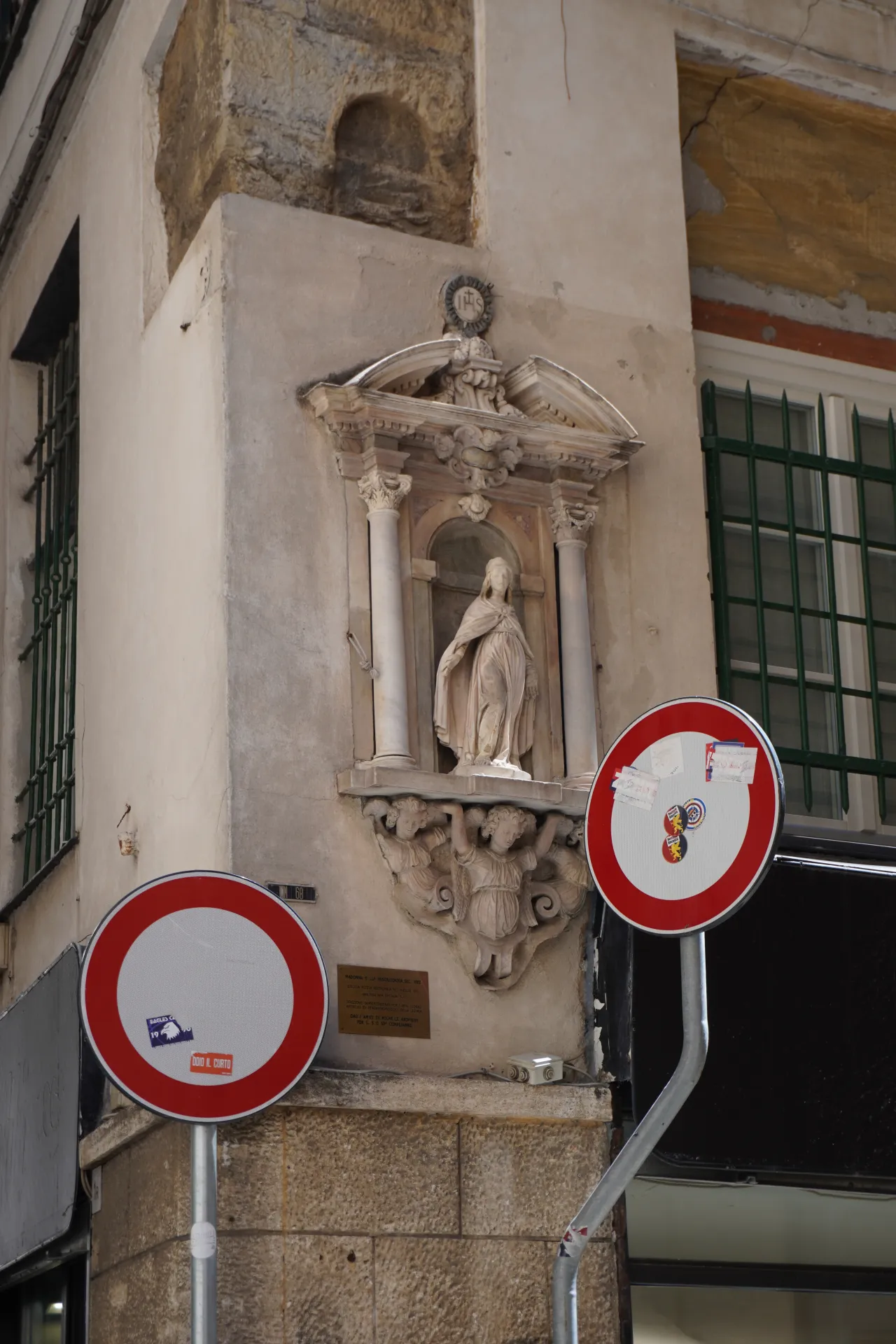 A religious niche with a statue of a woman is mounted on a building in Genoa, Italy, with two circular red-bordered street signs in the foreground.