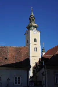 A tall, yellow bell tower with green and gold ornamentation and a cross on top, surrounded by terracotta tiled roofs, against a clear blue sky in Zagreb, Croatia.
