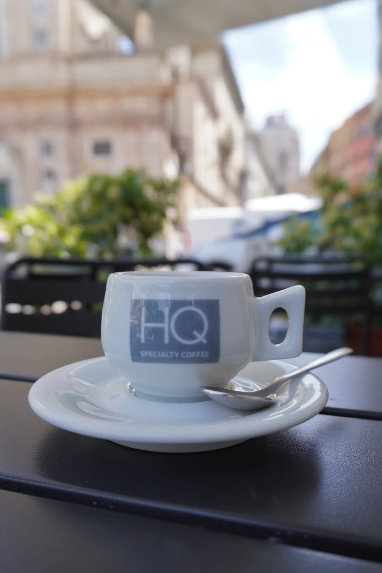 A white espresso cup with the 'HQ Specialty Coffee' logo sits on a saucer with a spoon, at an outdoor table in Genoa, Italy, with blurred buildings and greenery in the background.