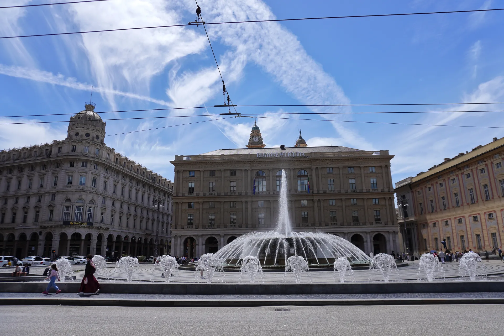 A large, circular fountain sprays water in the center of a piazza, surrounded by elegant historic buildings in Genoa, Italy. The sky above is blue with dramatic wispy clouds.
