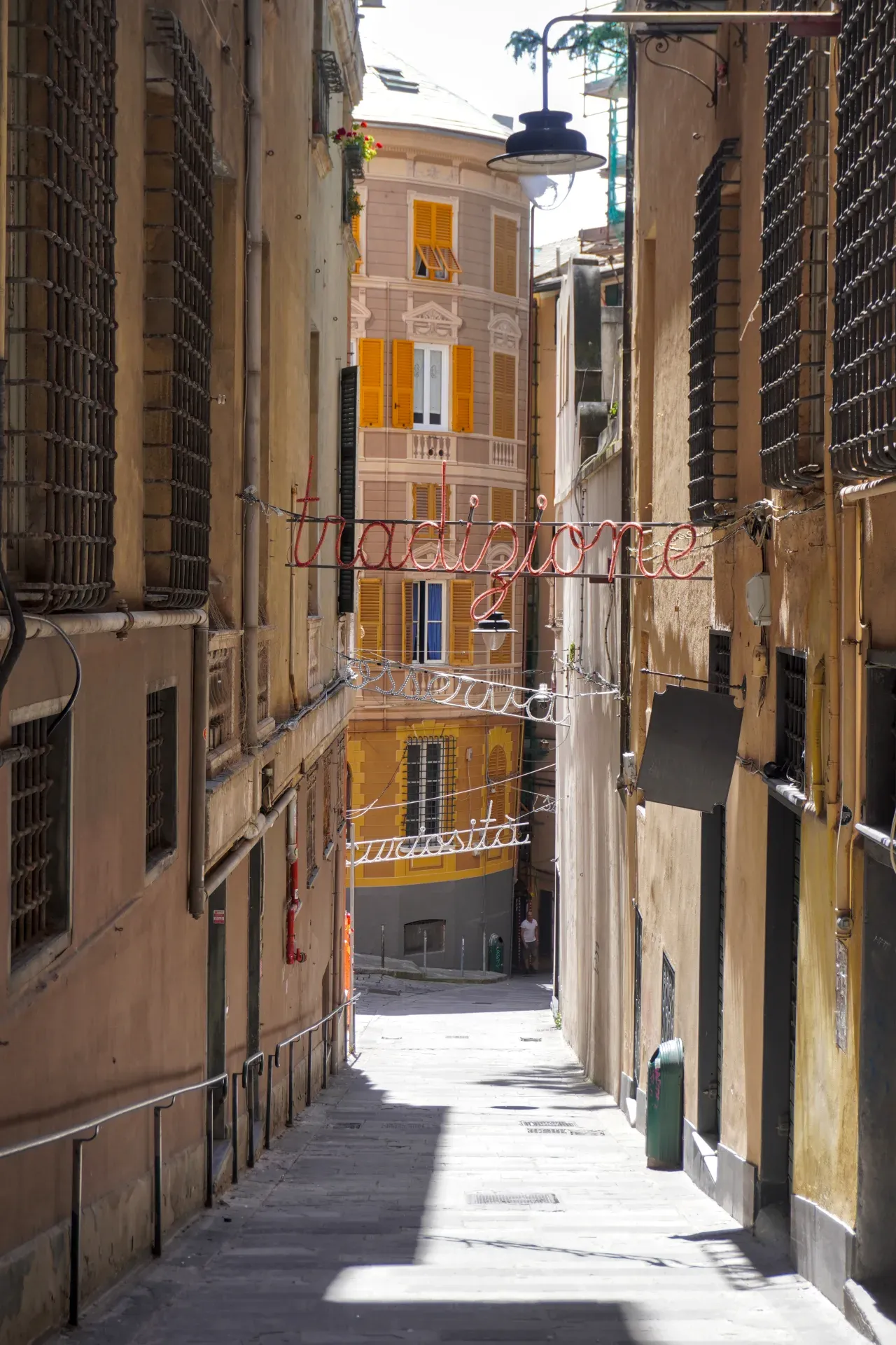 A narrow street in Genoa, Italy, with tall buildings on both sides and neon signs reading 'Tradizione' and 'Curiosità' hanging over the cobblestone pavement.