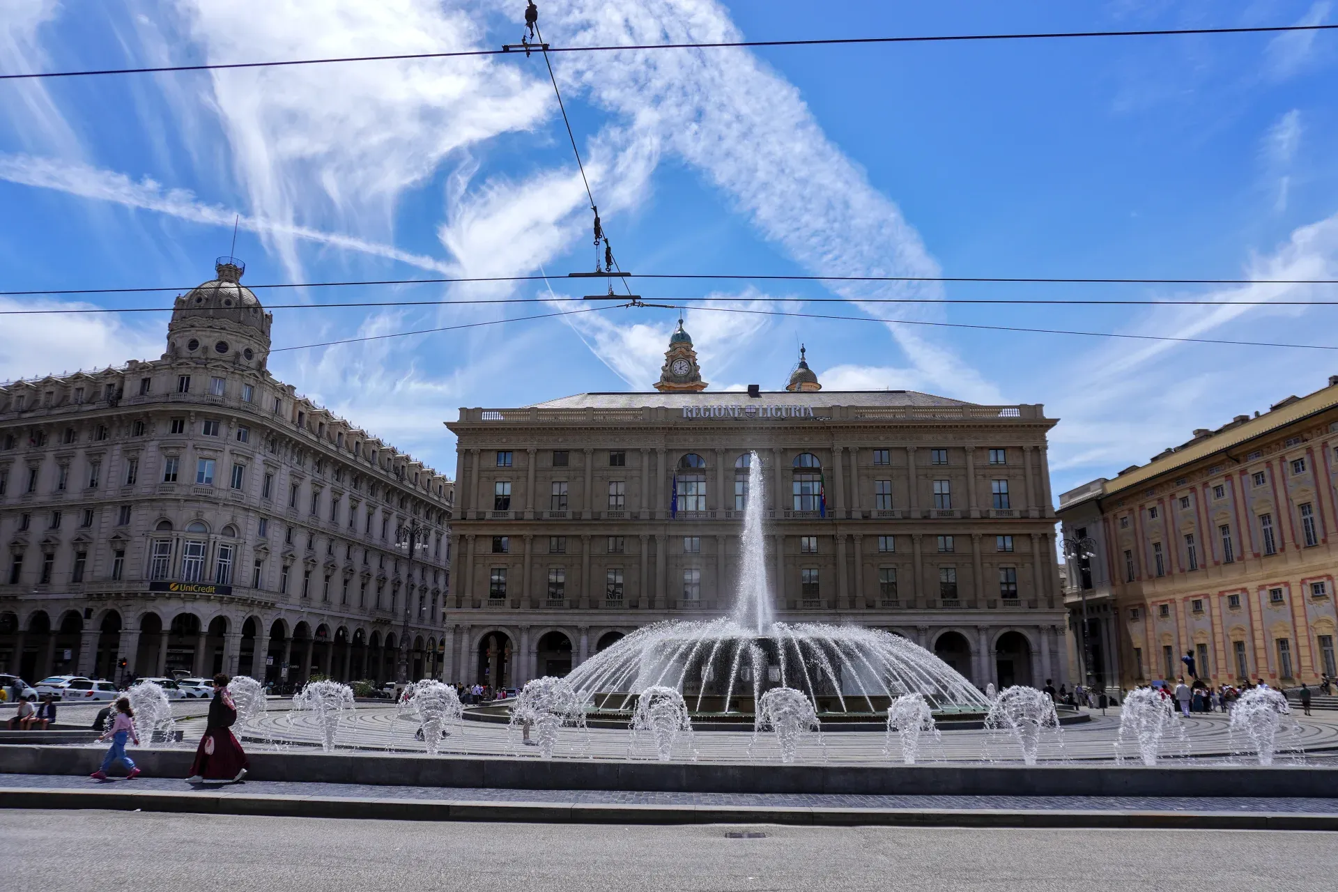 Velika fontana na više nivoa prska vodu na Piazza De Ferrari, Đenova, Italija. U prvom planu su beli automobili, jedan je taksi, i biciklista na putu. Iza fontane je raskošna žuta zgrada sa ružičastim detaljima ispod plavog neba sa belim oblacima.