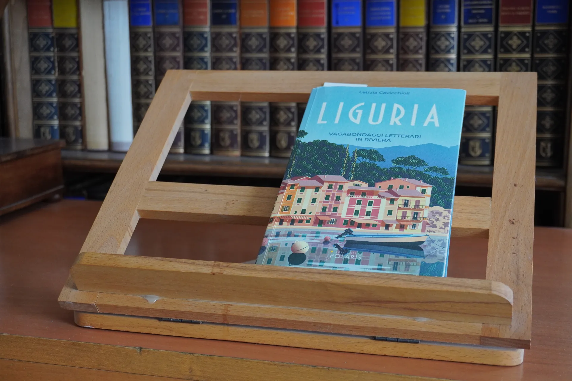 Knjiga sa šarenom ilustracijom ligurijskih primorskih kuća i čamcem na drvenom postolju, sa policom starih knjiga u pozadini.