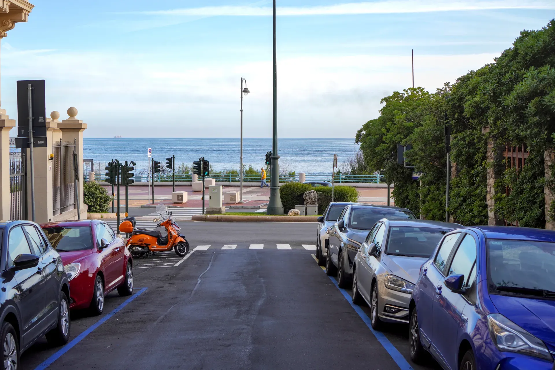 A street in Genoa, Italy, with cars parked along the sides and the sea in the background, framed by buildings and trees.