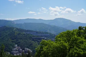 View of Genoa, Italy, with buildings integrated into verdant, rolling hills under a blue sky with scattered clouds.