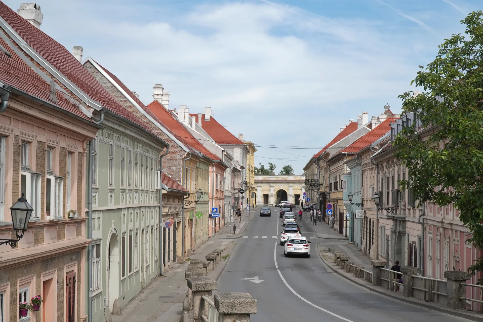 Вид вниз по улице в Нови-Саде, Сербия, с красочными историческими зданиями по обеим сторонам, автомобилями, движущимися по дороге, и аркой вдалеке под частично облачным небом.