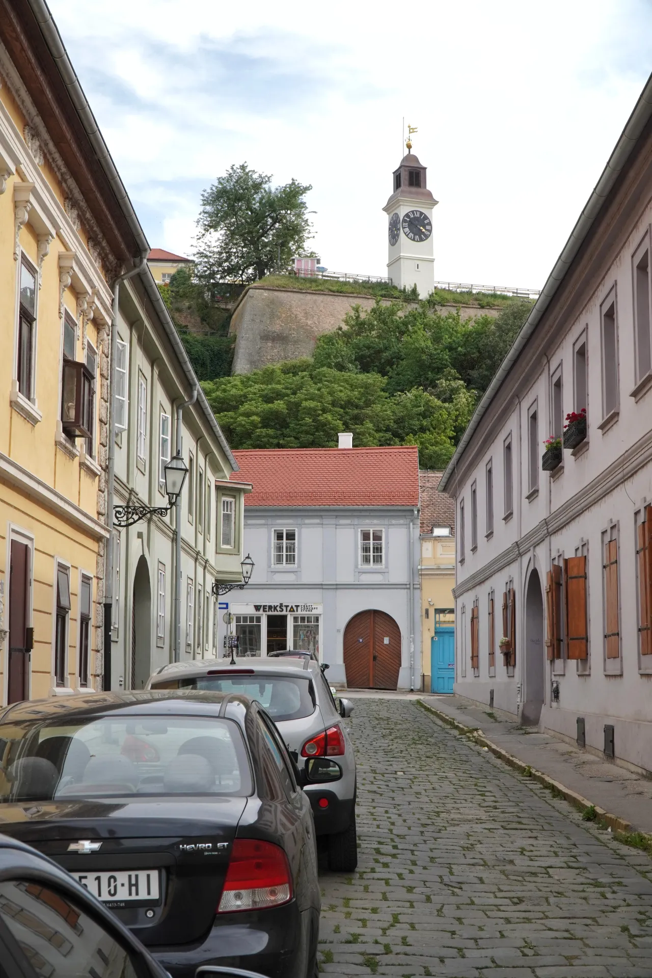 A cobblestone street in Novi Sad, Serbia, lined with colorful buildings, leading to the Petrovaradin Fortress clock tower visible in the background.