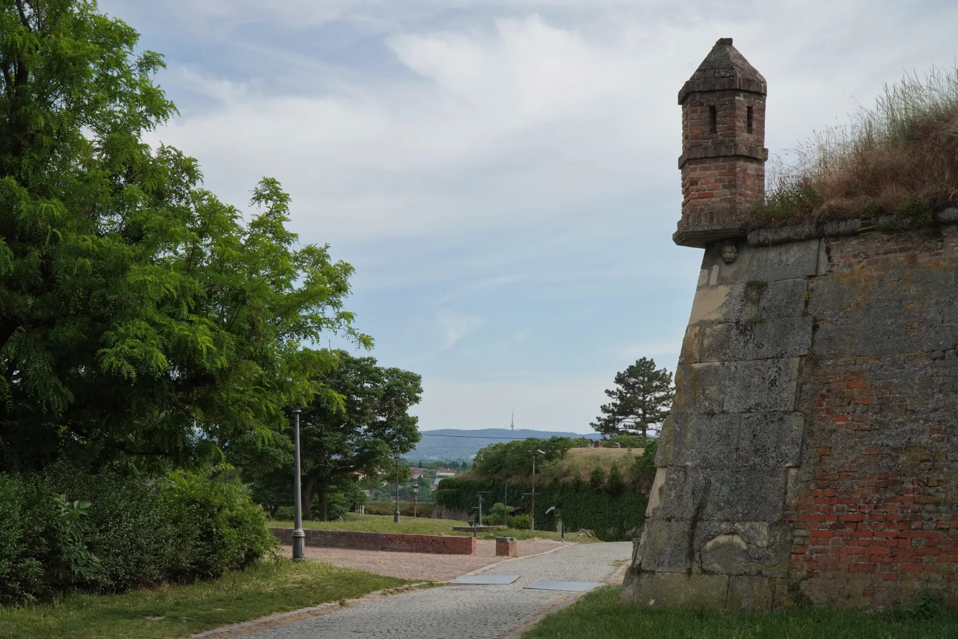 A detailed view of an old brick watchtower on the stone walls of Petrovaradin Fortress in Novi Sad, Serbia, with trees and a path in the foreground and a hazy landscape in the background.