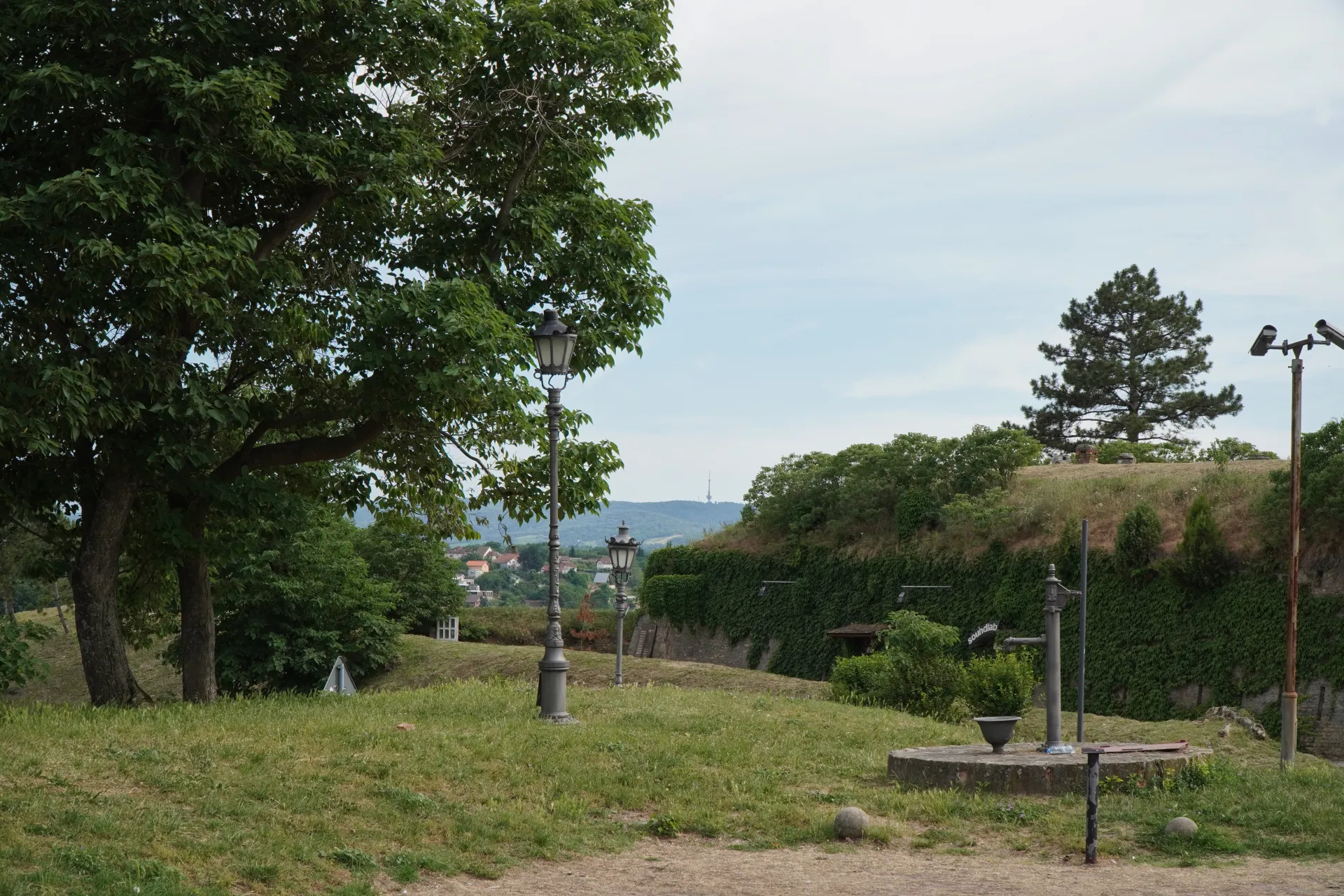A view from Petrovaradin Fortress with trees, street lamps, an old water pump, and the city of Novi Sad in the background.