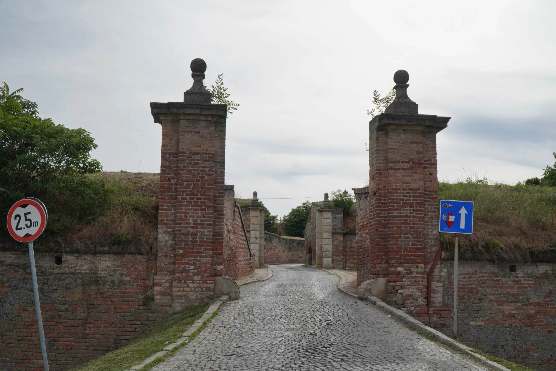 Мощеная дорога, ведущая через кирпичные ворота Петроварадинской крепости, Нови-Сад, Сербия, с травянистыми валами и пасмурным небом.