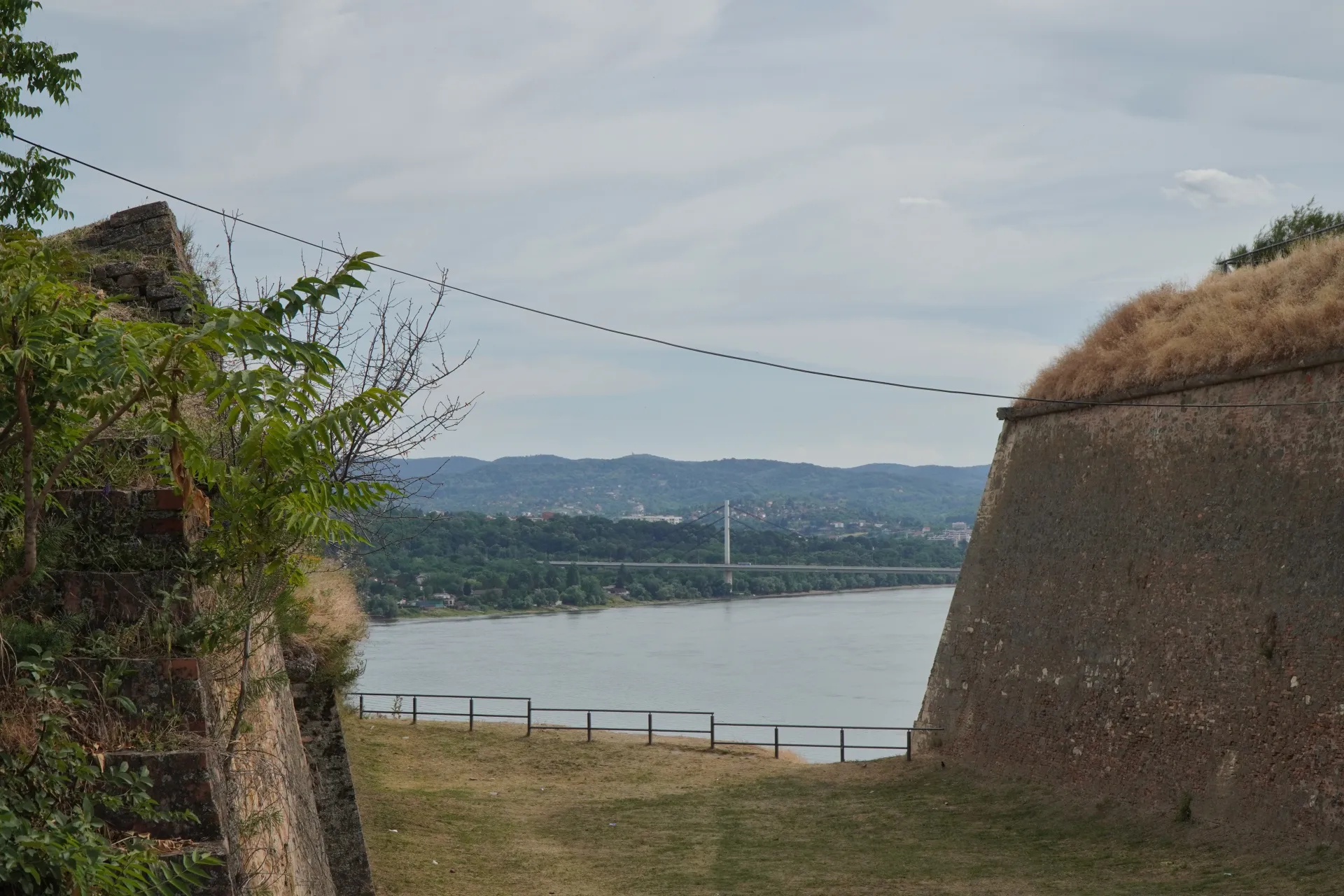 View of the Danube River and Novi Sad from the historic Petrovaradin Fortress, featuring old stone walls, vegetation, and a distant bridge.