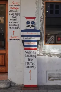A white sign with black lettering listing "SMALL ANTIQUES", "VINTAGE", "WATCHES", "SUNGLASSES", "PENS", "FLUTES", "DOC BAGS", "LIGHTERS", and "FIGURINES", with an orange arrow pointing upwards, stands next to a stylized wooden figure in front of a shop in Zagreb, Croatia.