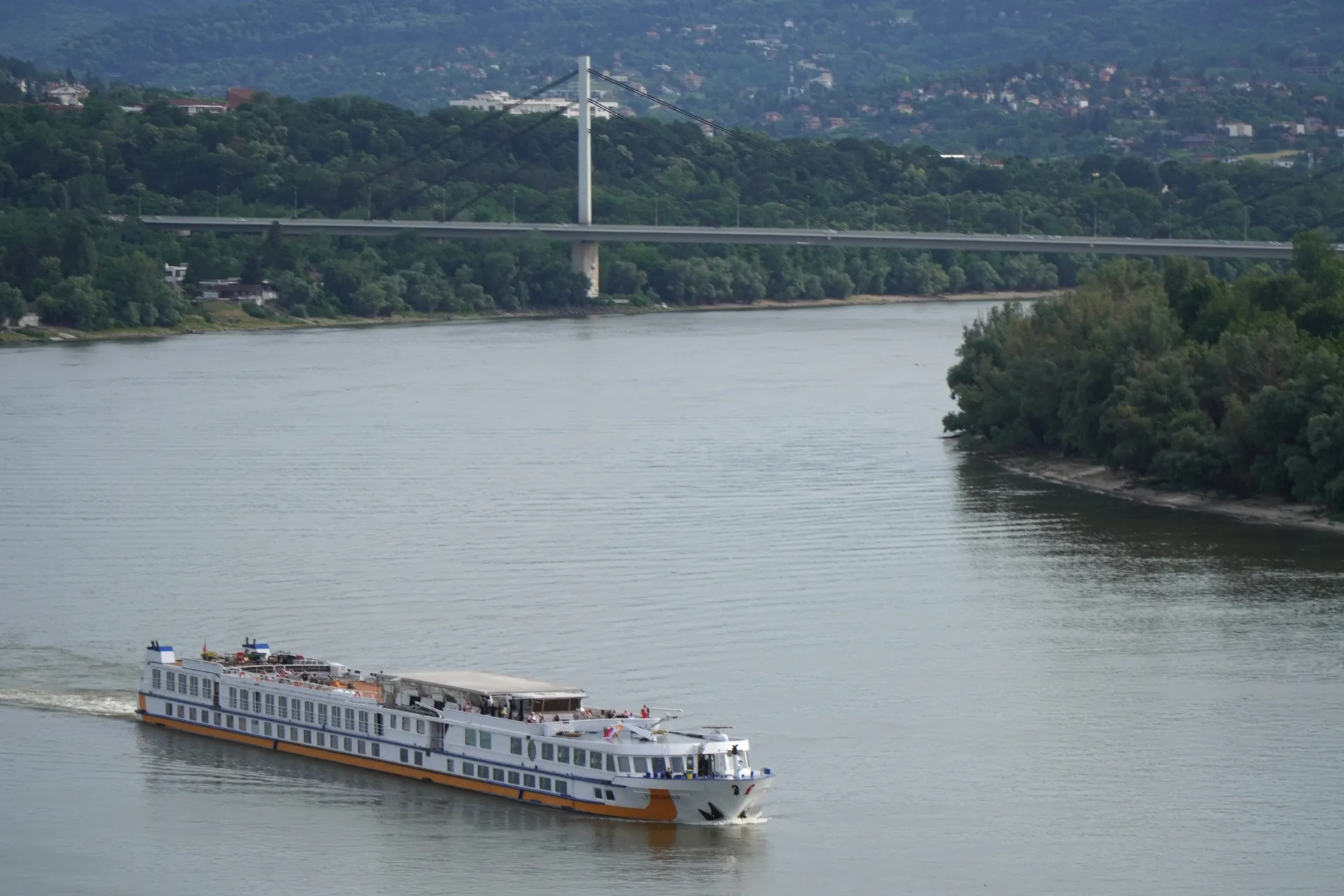 Beli i narandžasti brod za krstarenje plovi Dunavom, sa dugim modernim mostom koji preseca vodu u pozadini. Obala je prekrivena bujnim zelenim drvećem, a u daljini se vide brda sa zgradama, što ukazuje na lokaciju u blizini Novog Sada, Srbija.