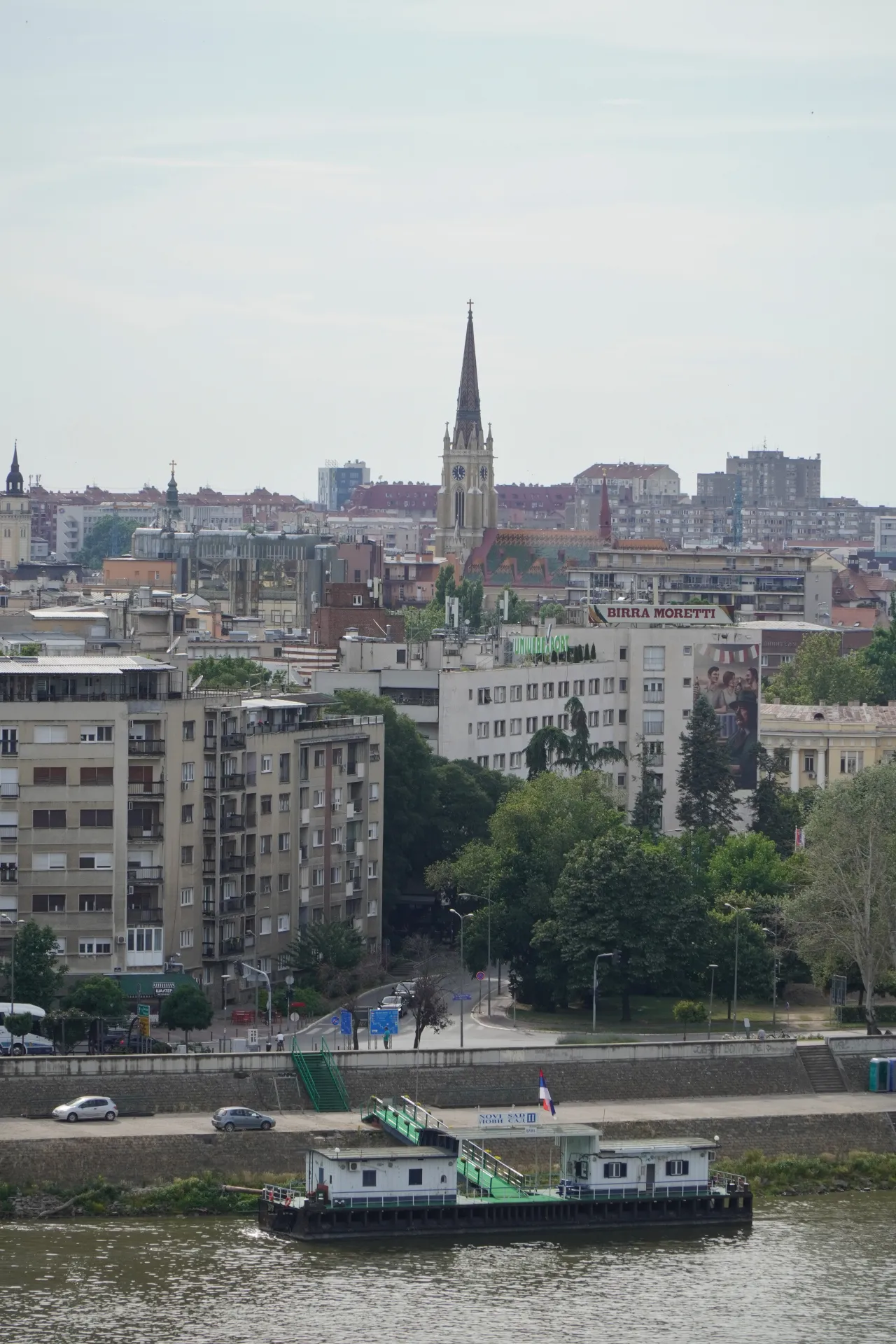 A wide shot of the Novi Sad cityscape overlooking the Danube River. A church spire rises above the buildings, and a barge is docked by the riverside.